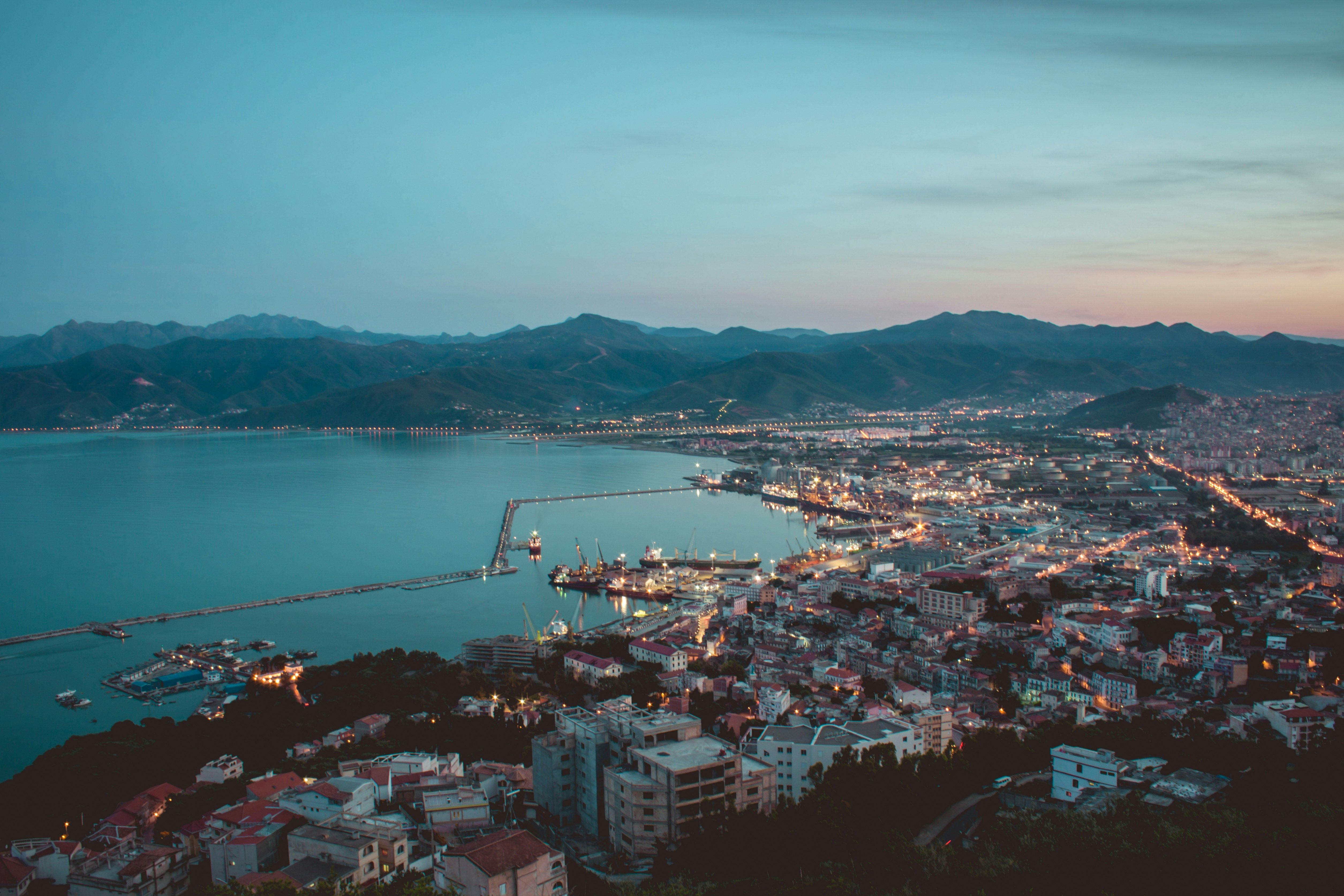 Vue plongeante au crépuscule sur une ville côtière illuminée, un port et ses longues jetées ; au fond, une baie bordée de montagnes
