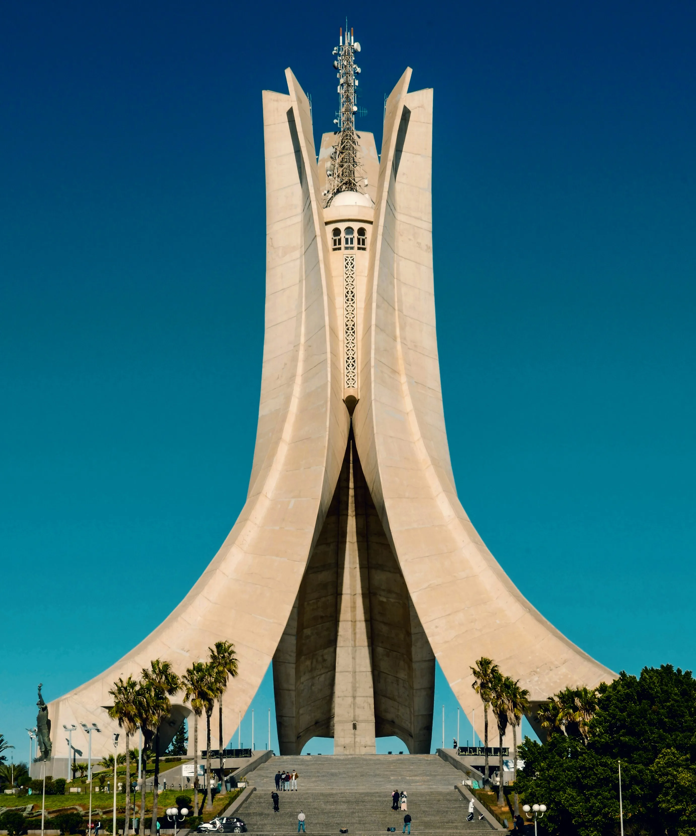 Monument de béton clair aux grandes nervures qui s’élèvent et se resserrent autour d’une tourelle, au sommet d’un large escalier bordé de palmiers