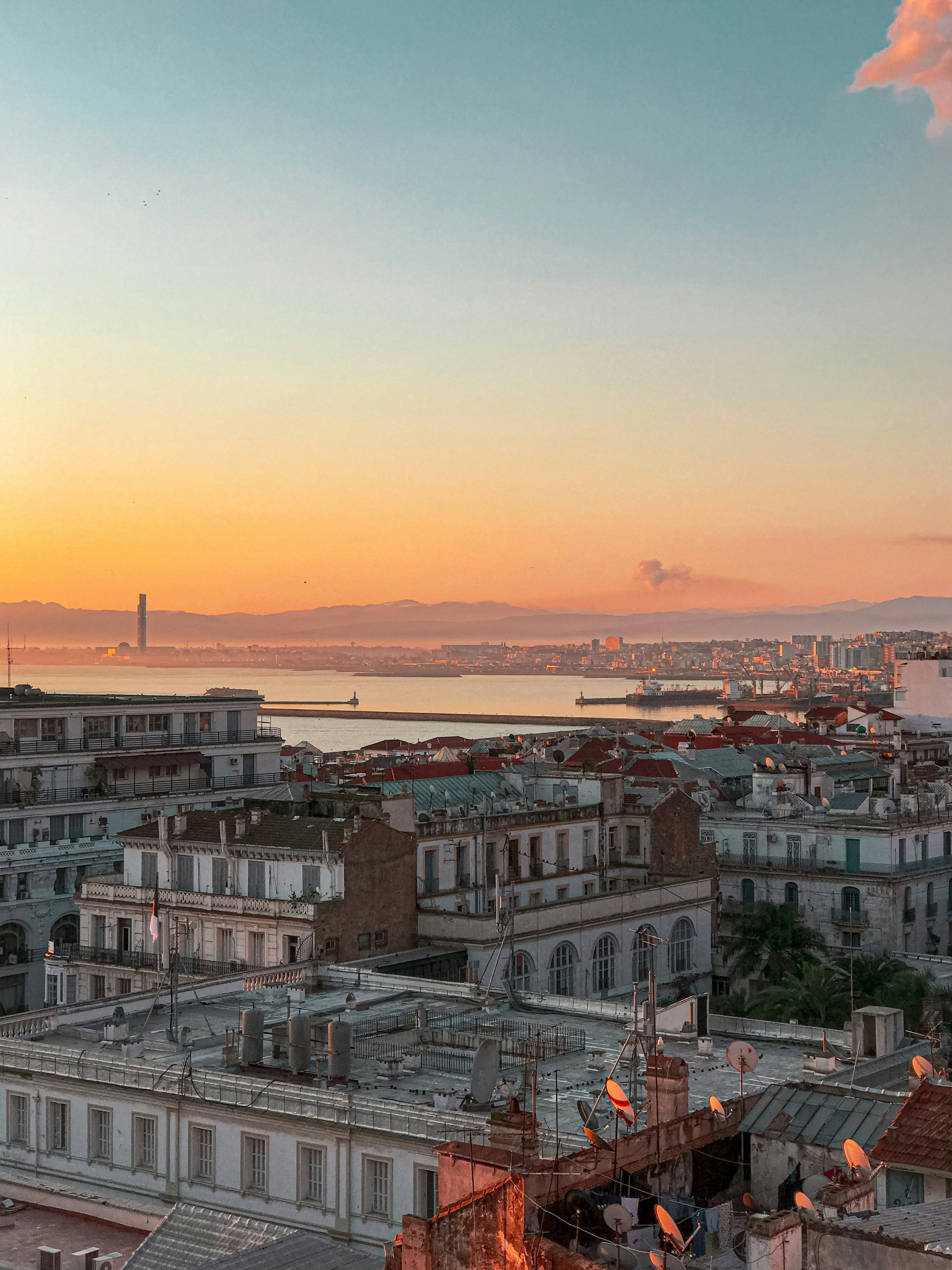 Toits en terrasse et immeubles clairs au premier plan, un port fermé par une jetée, puis la baie et des montagnes à l’horizon, sous un ciel orangé