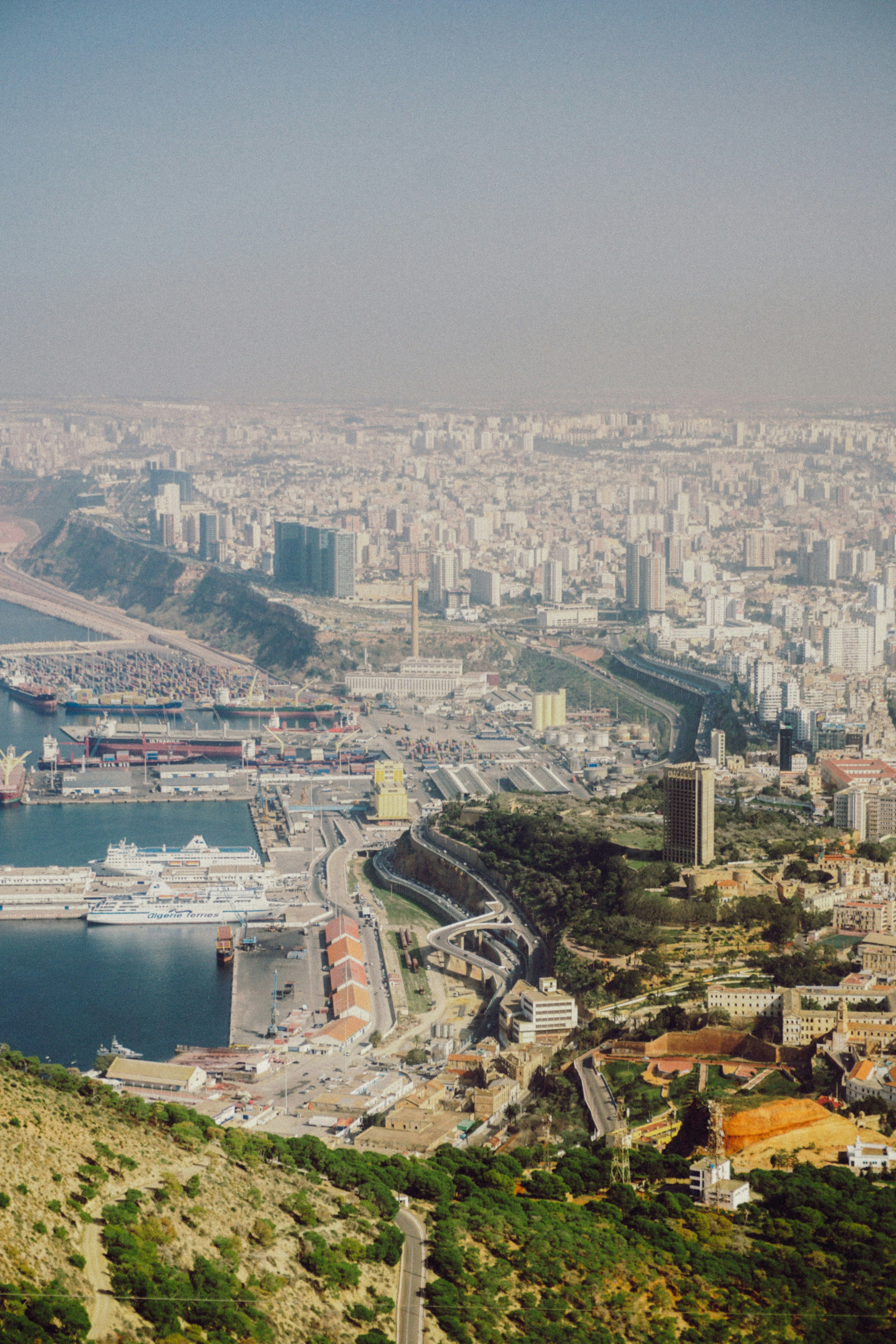 Vue plongeante sur une ville dense qui s’étend jusqu’à l’horizon brumeux ; au pied d’une colline boisée, un port où sont amarrés deux grands navires blancs, et une voie rapide qui serpente entre les quais et la ville