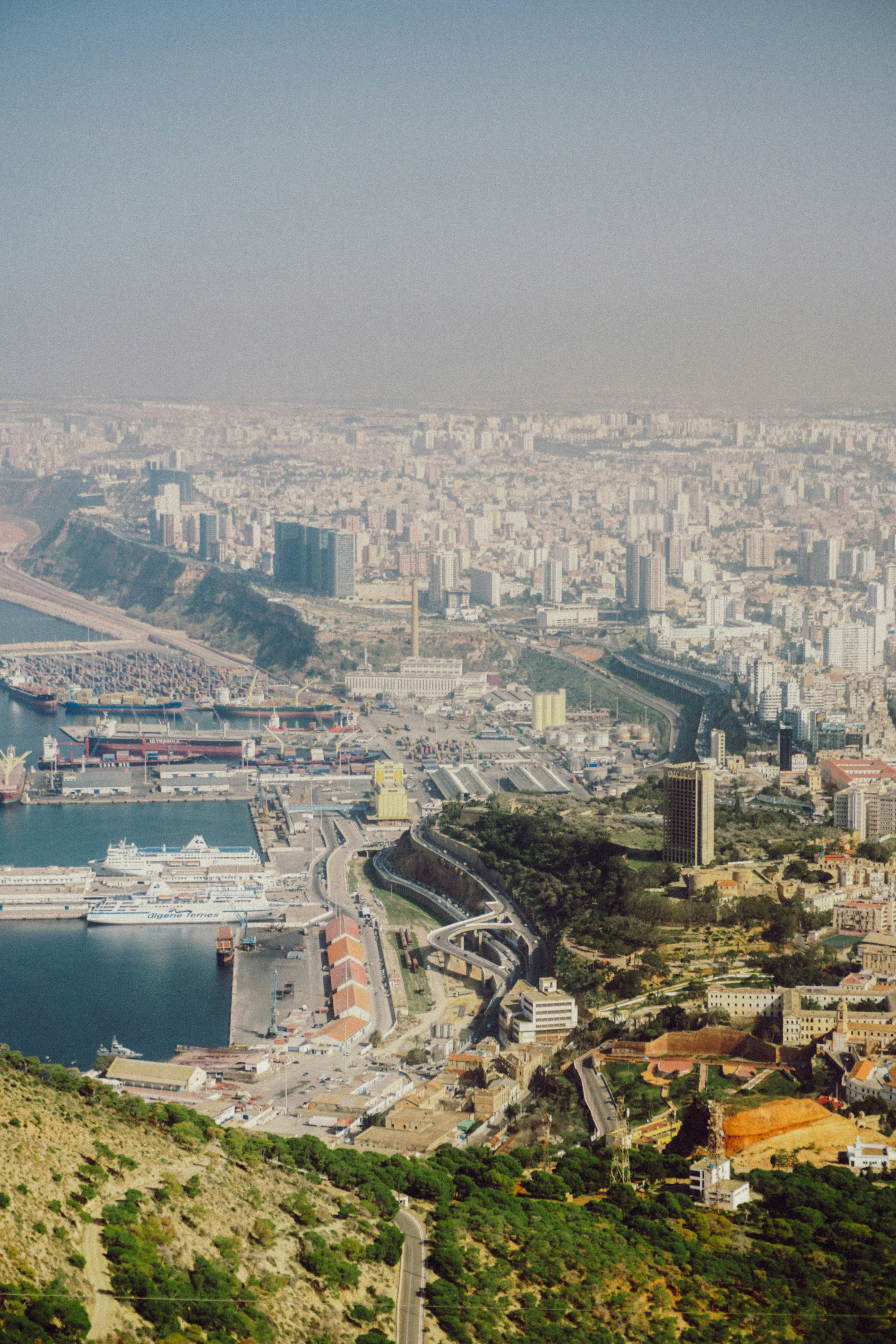 Vue plongeante sur une ville dense qui s’étend jusqu’à l’horizon brumeux ; au pied d’une colline boisée, un port où sont amarrés deux grands navires blancs, et une voie rapide qui serpente entre les quais et la ville