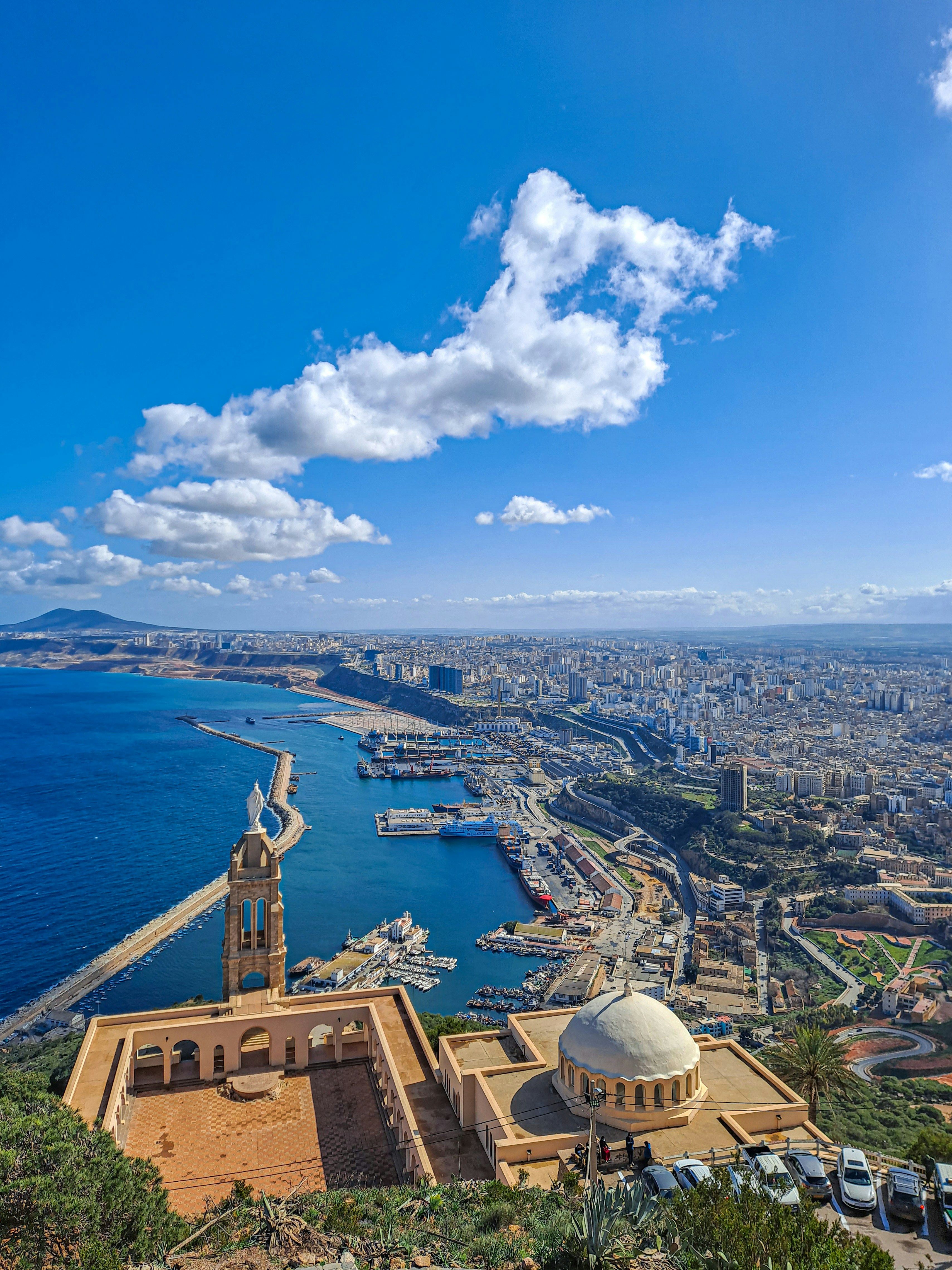 Vue plongeante depuis une colline : une tour de pierre surmontée d’une statue blanche, une esplanade bordée d’arcades et une coupole blanche, avec le port et la ville en contrebas
