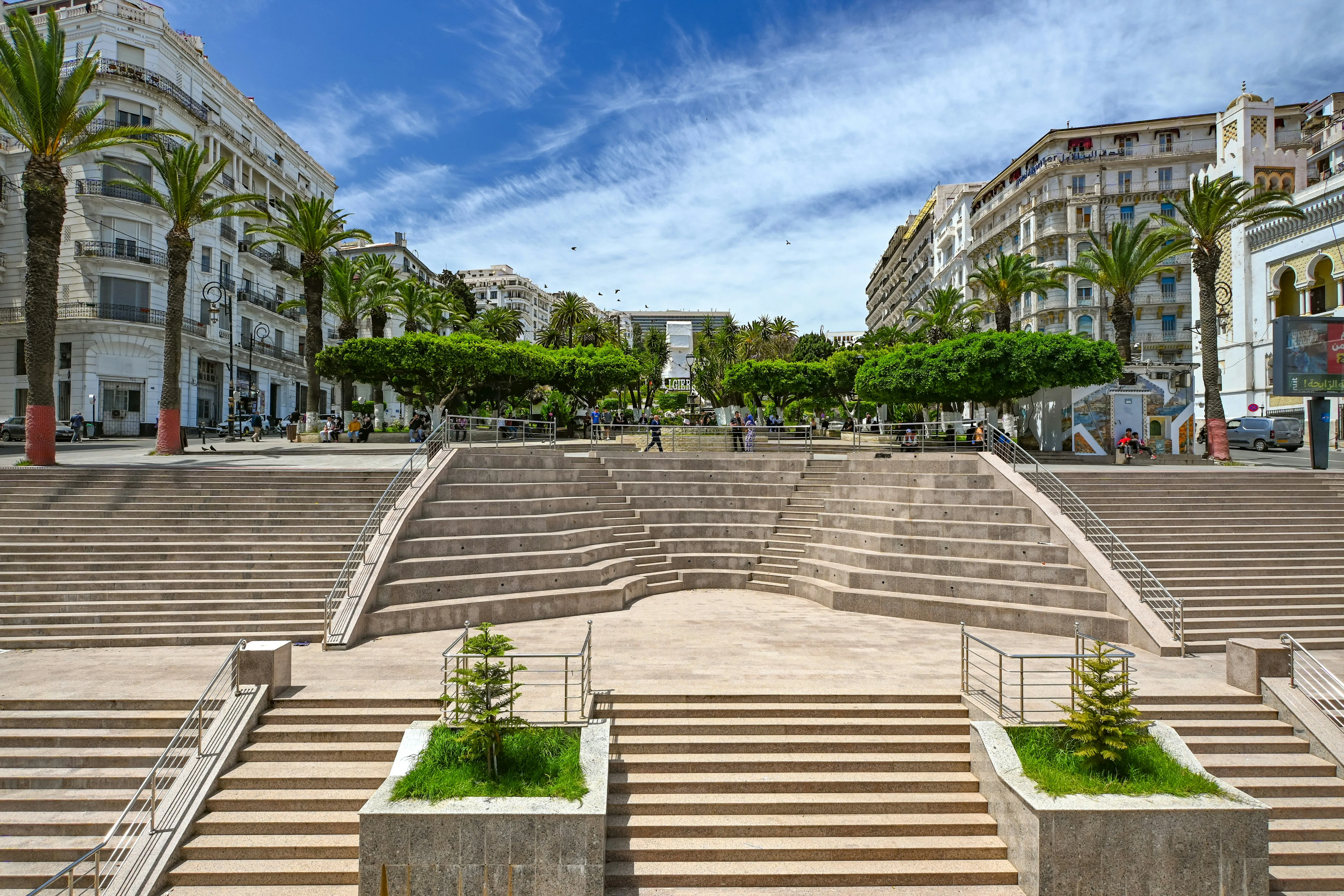 Grand escalier de pierre claire en gradins, entre des immeubles blancs à balcons et des palmiers ; en haut, une place arborée et, à droite, une façade blanche à arcs outrepassés