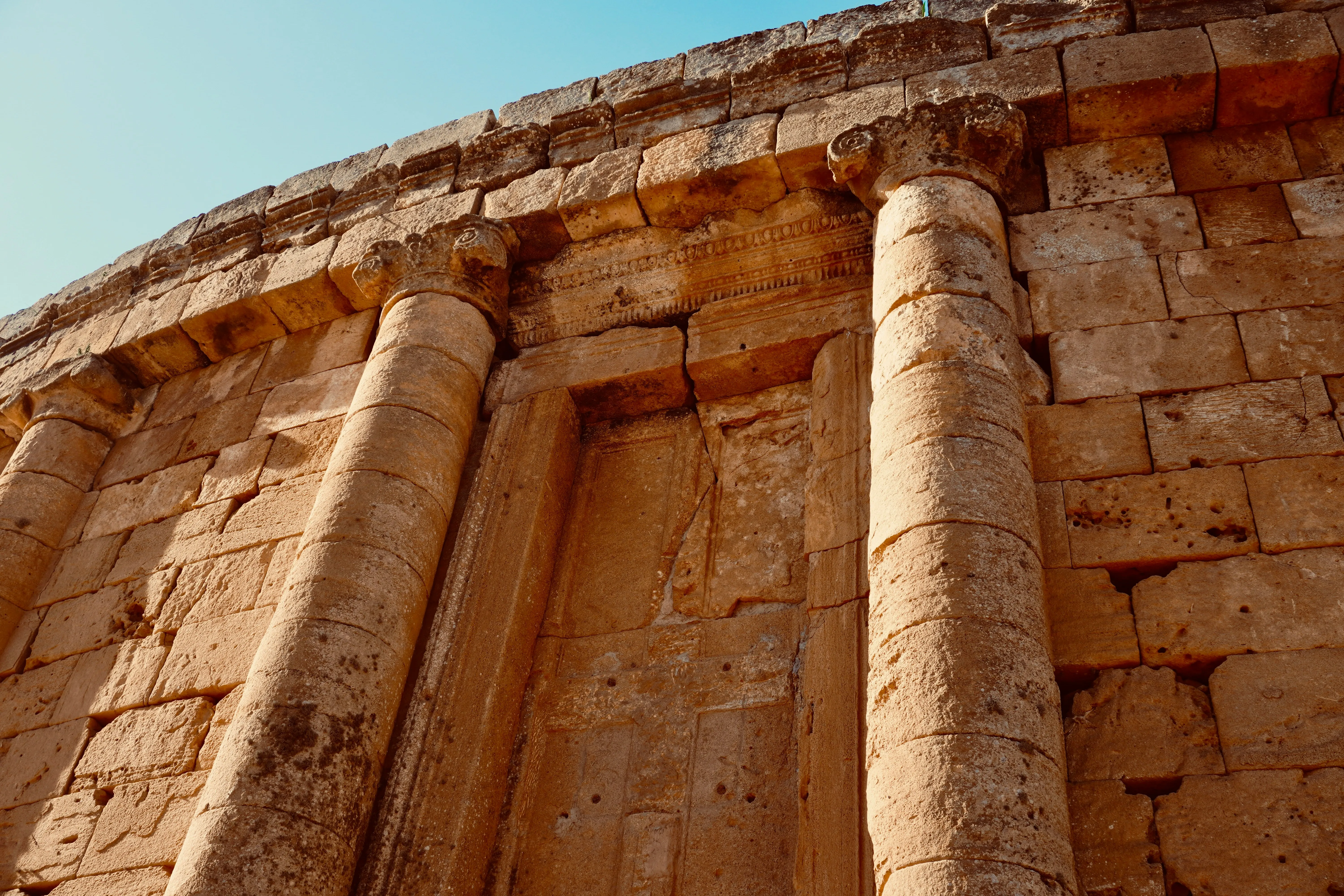 Mur circulaire en gros blocs de pierre ocre, vu d’en bas ; deux colonnes engagées à chapiteaux encadrent une fausse porte surmontée d’une frise sculptée