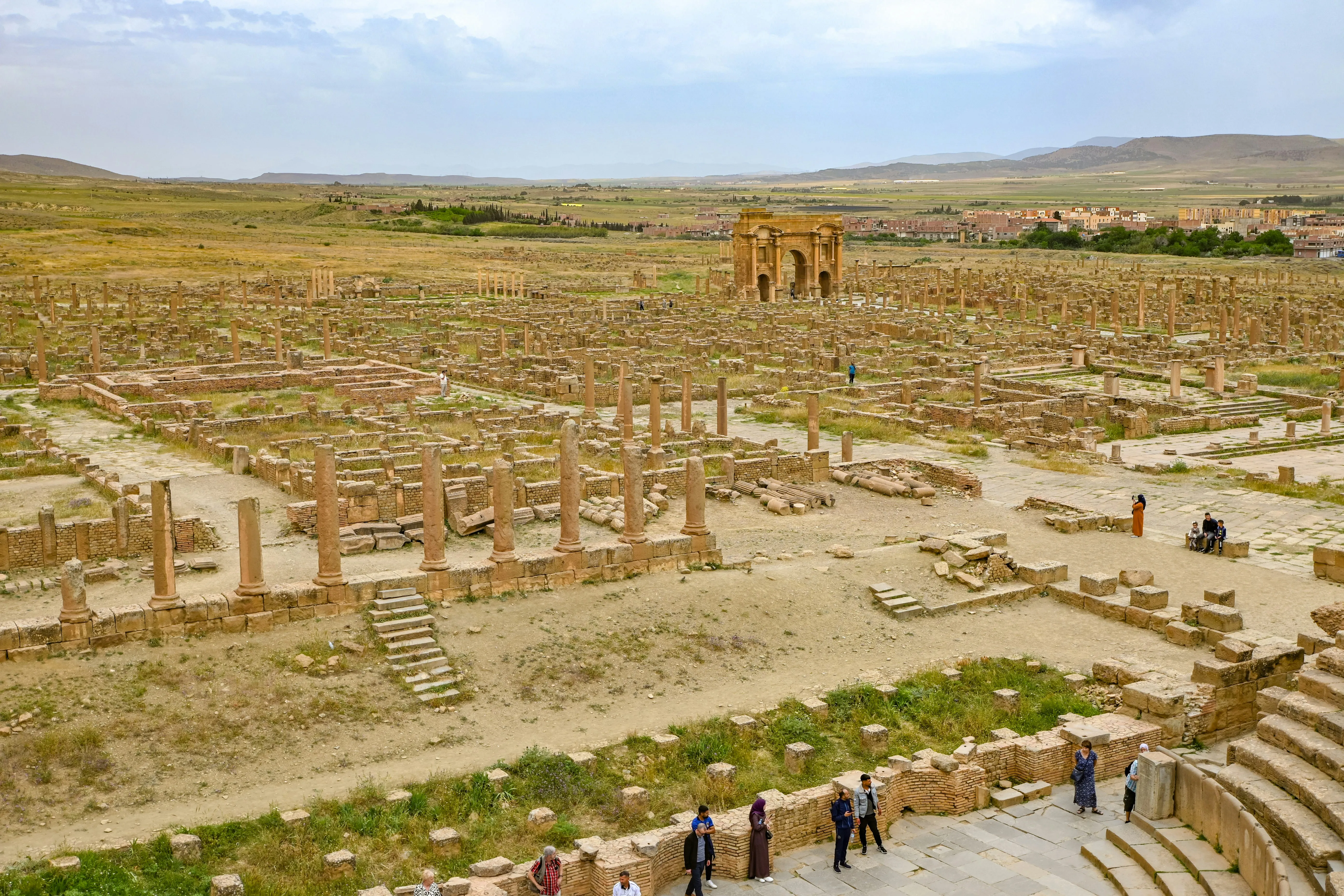 Champ de ruines ocre à perte de vue : colonnes dressées, murets et rues en damier, avec au centre un grand arc à trois baies ; au fond, une ville basse, des champs et des montagnes sous un ciel nuageux