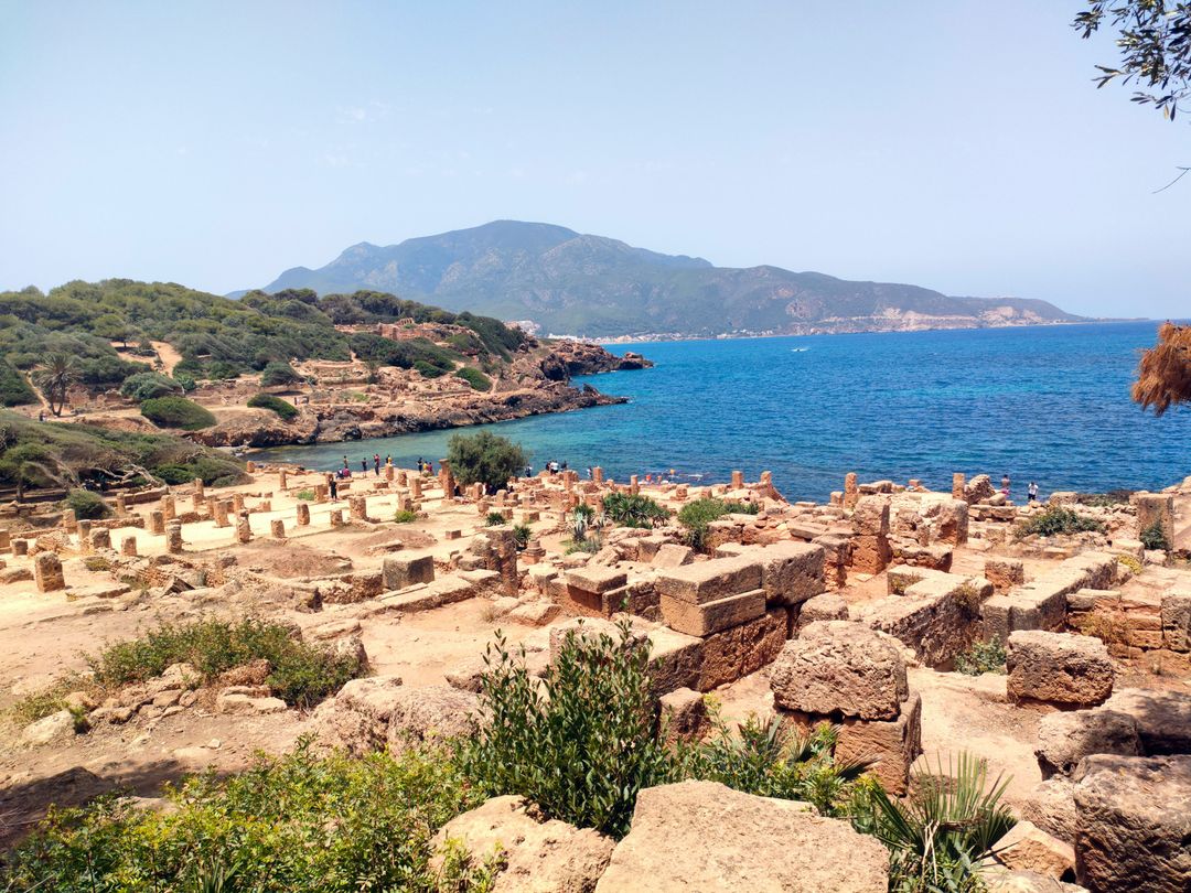 Champ de ruines en pierre ocre, colonnes tronquées et murets, au bord d’une crique bleue ; au fond, une montagne boisée domine la mer