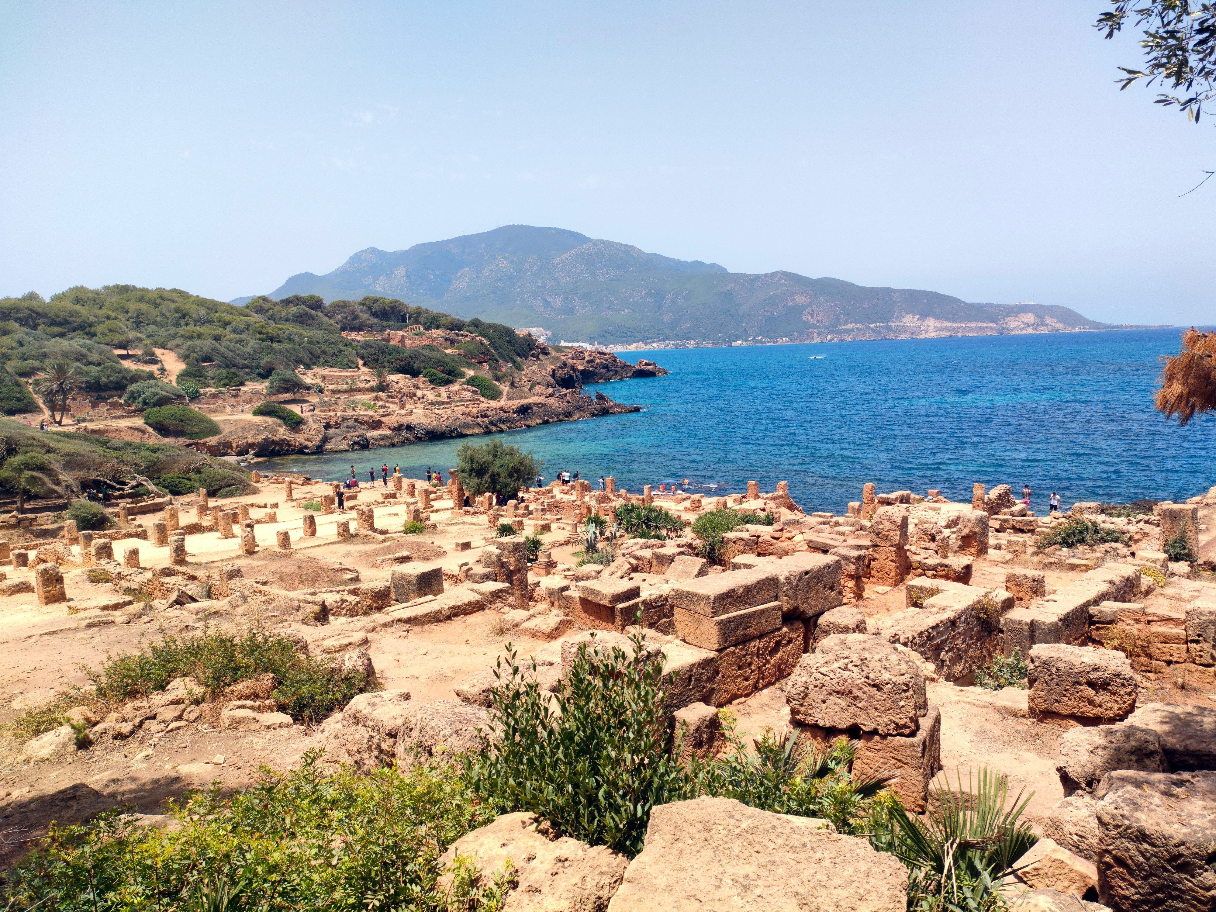 Champ de ruines en pierre ocre, colonnes tronquées et murets, au bord d’une crique bleue ; au fond, une montagne boisée domine la mer