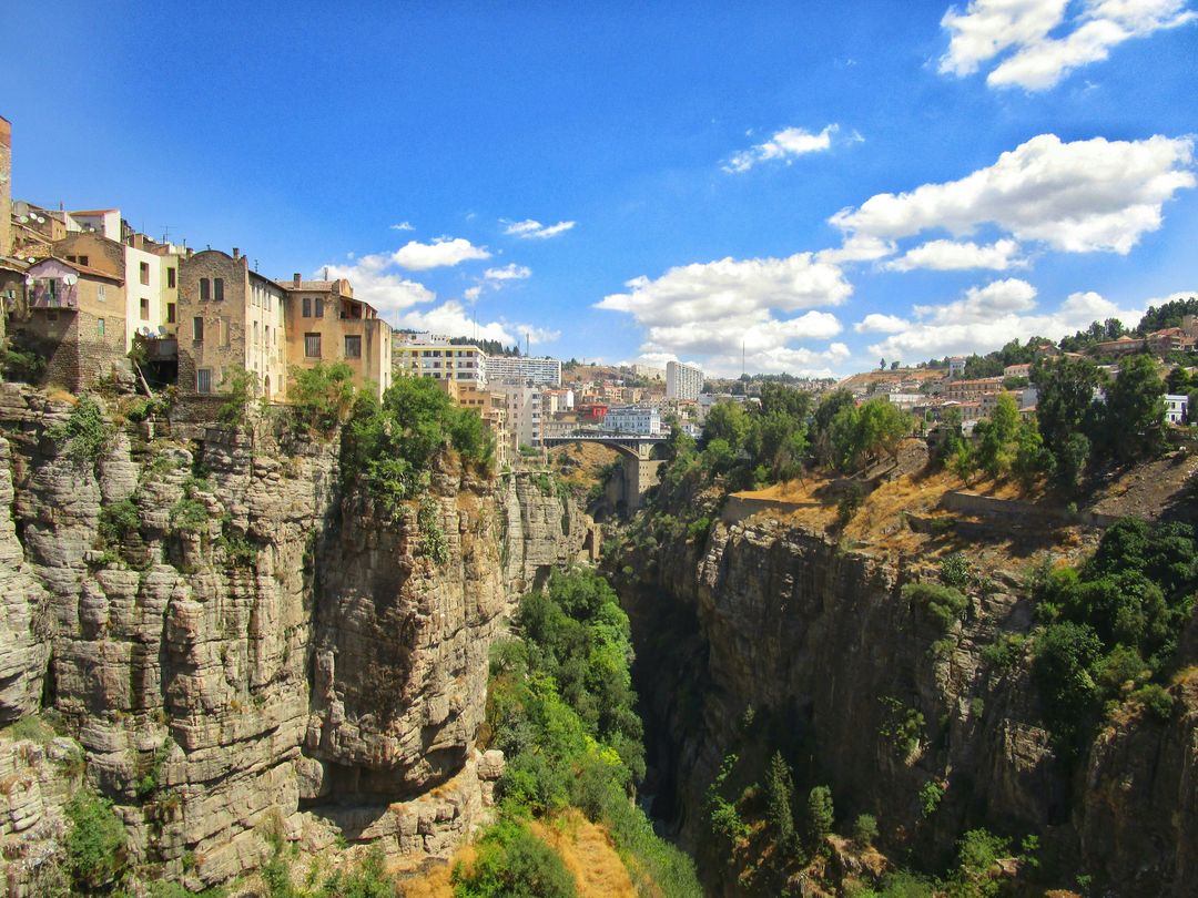Maisons de plusieurs étages alignées au bord d’une falaise calcaire, au-dessus d’une gorge étroite et boisée ; au fond, un pont à arches et des immeubles sur la colline