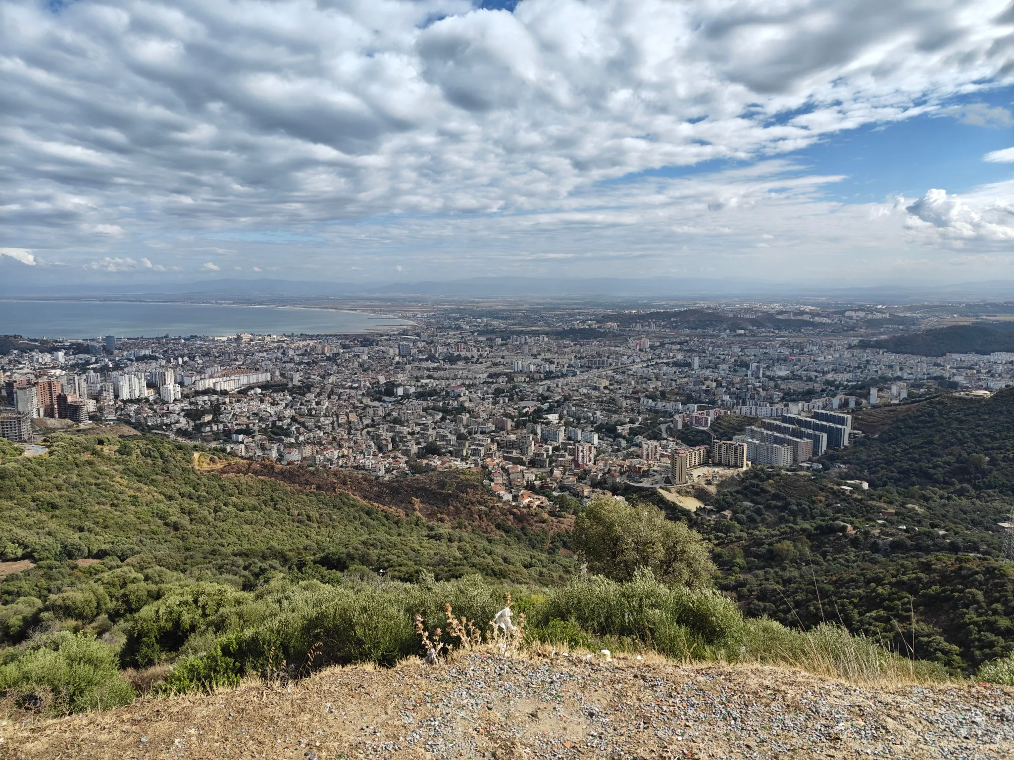 Vue plongeante sur une grande ville blanche qui s’étend jusqu’à une baie, sous un ciel chargé de nuages ; au premier plan, des collines couvertes de maquis
