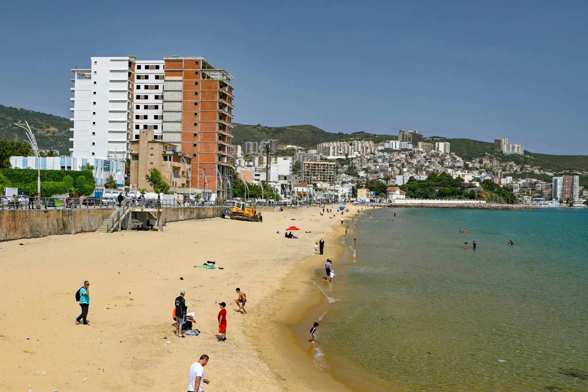 Plage de sable bordée d’une promenade et d’immeubles, avec des promeneurs sur le sable ; au fond, la ville s’étage sur des collines vertes