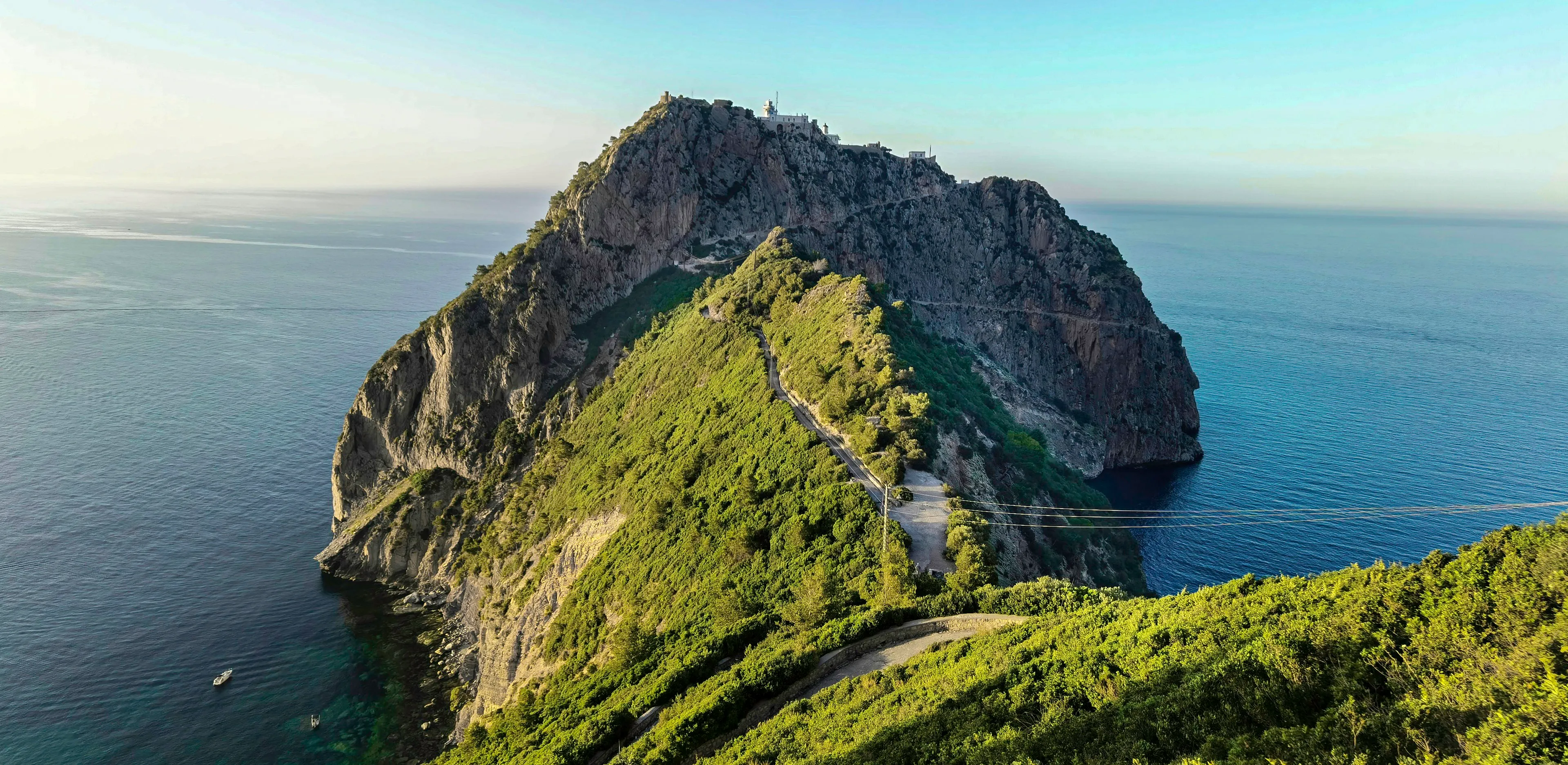 Presqu’île rocheuse couverte de végétation, entourée par la mer ; une route en lacets monte vers un bâtiment clair au sommet