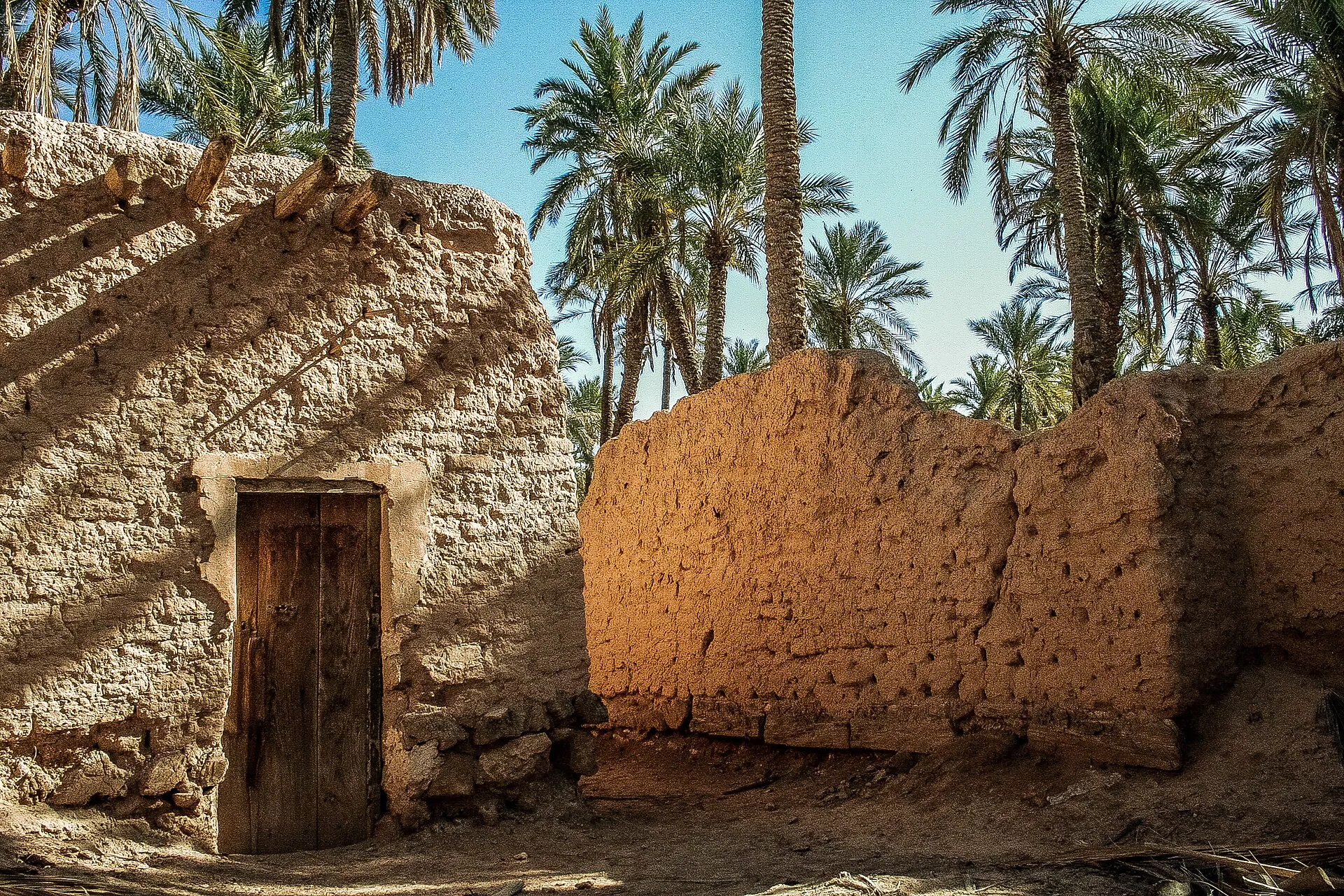 Une petite maison aux murs de terre, avec une porte de bois sombre, à côté d’un haut mur de terre éclairé par le soleil ; des palmiers derrière