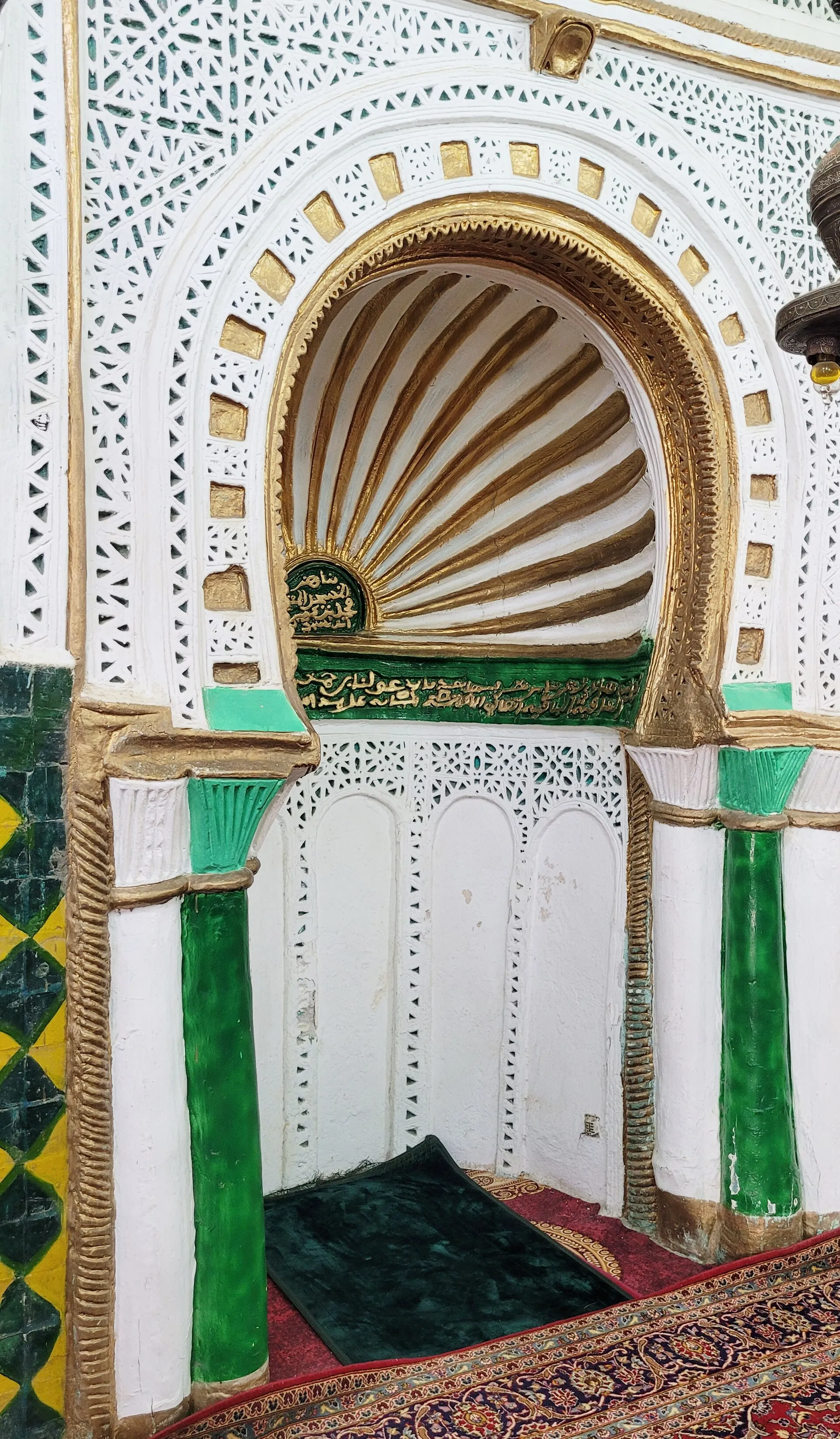 Une niche de prière en arc outrepassé, coiffée d’une coquille cannelée dorée, encadrée de stuc blanc ajouré et de deux colonnes vertes ; un tapis au sol