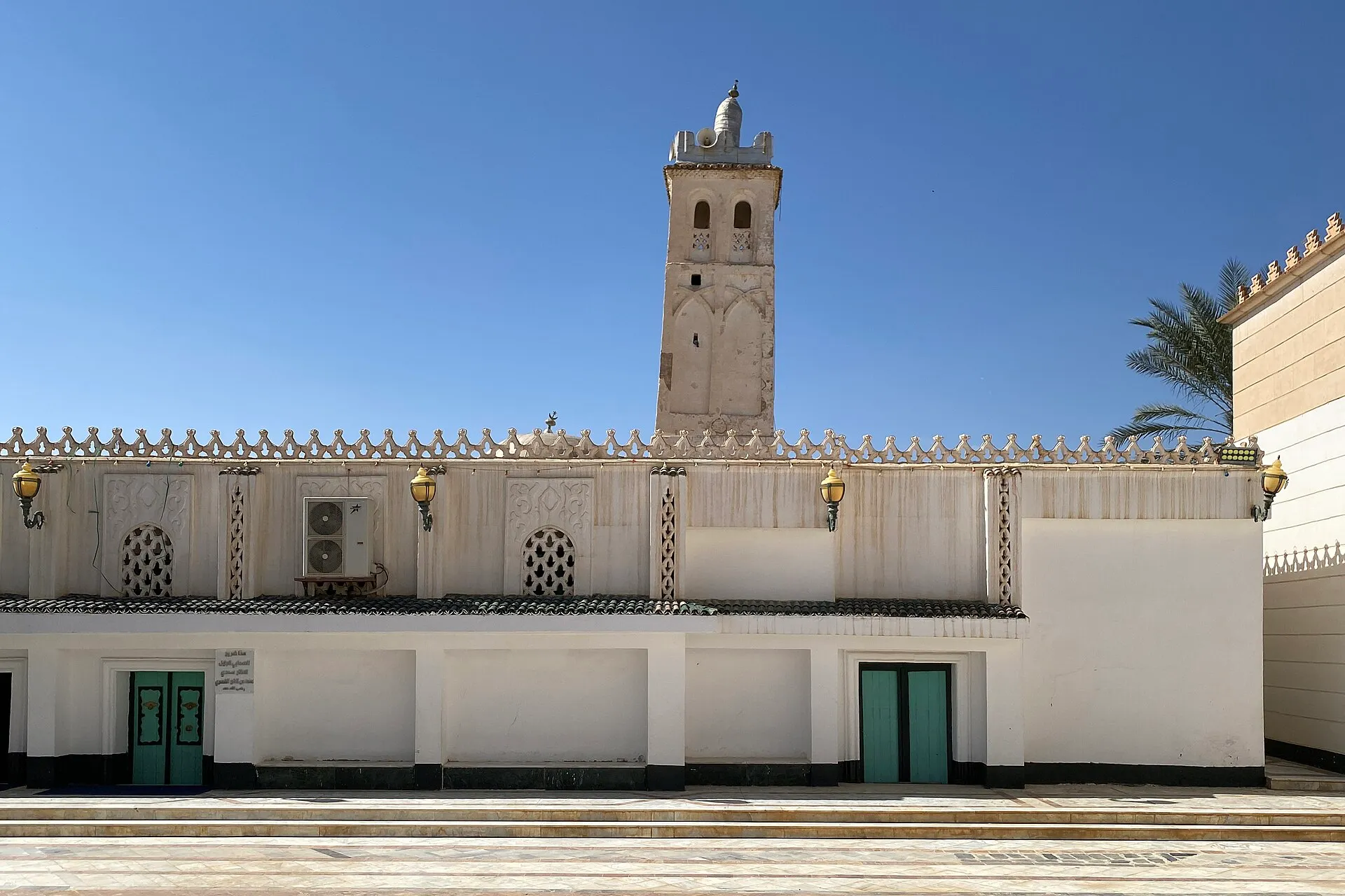 Un long mur blanc couronné de merlons dentelés, percé de portes vertes, derrière lequel s’élève un minaret carré à deux niveaux de baies, sous un ciel bleu