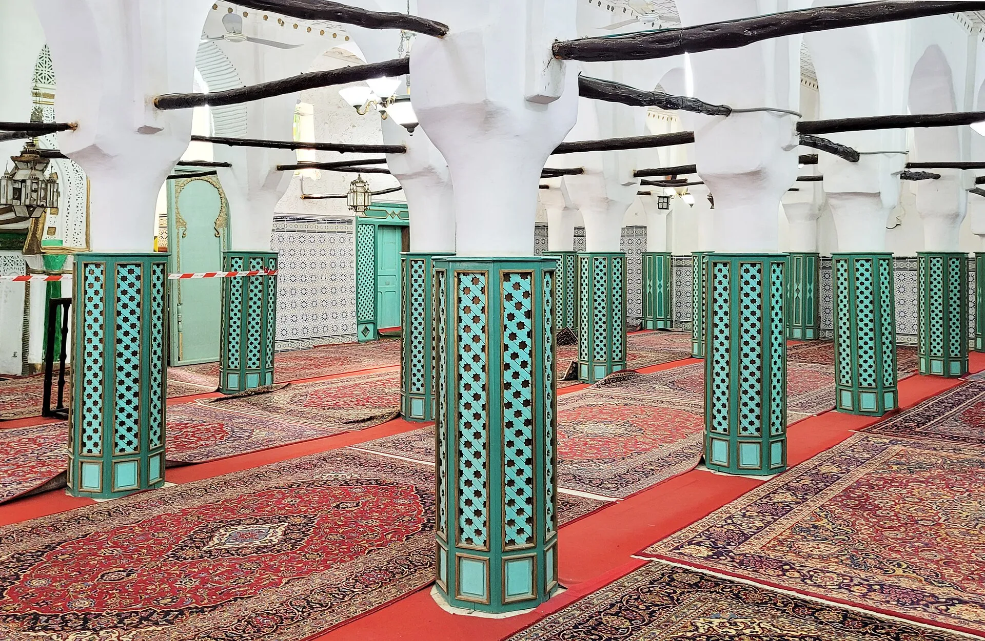 Une salle de prière aux piliers blancs évasés vers le haut, posés sur des socles octogonaux verts ajourés, sous des poutres sombres ; des tapis rouges couvrent le sol