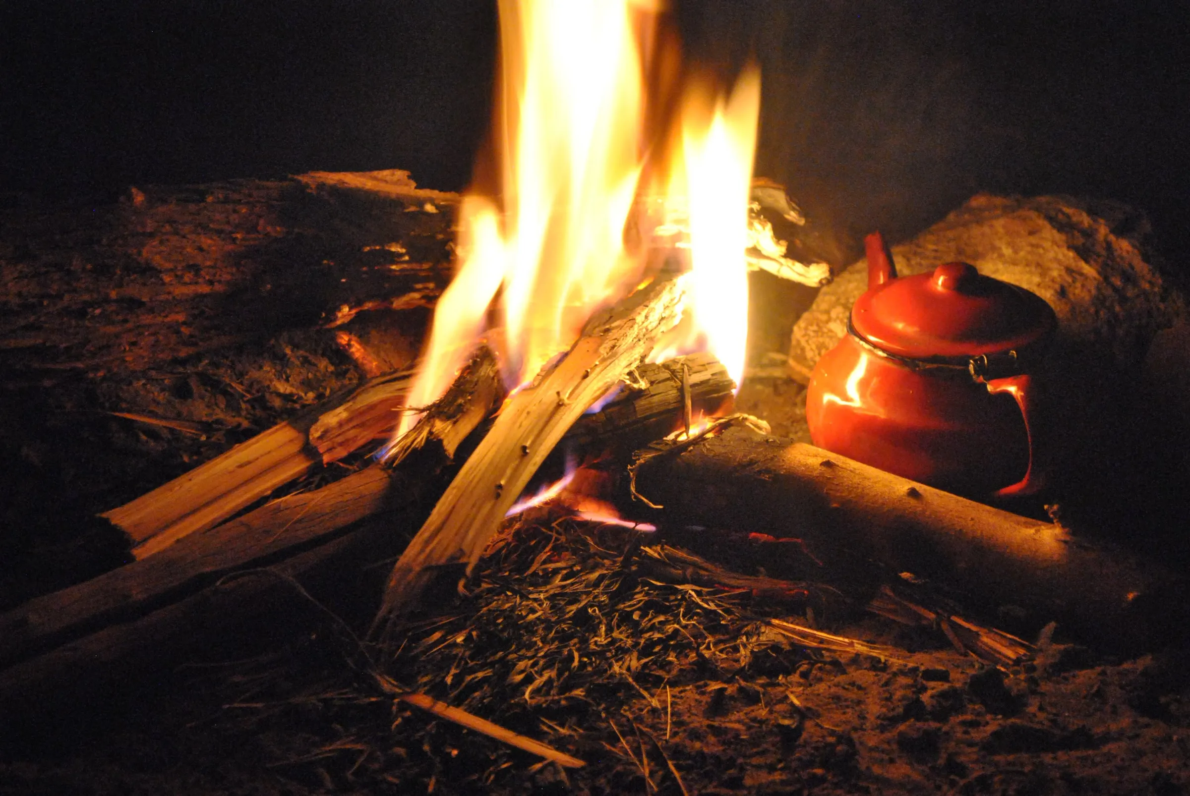 De nuit, un feu de bois vif dont les flammes éclairent une théière rouge émaillée posée dans les braises, sur le sable