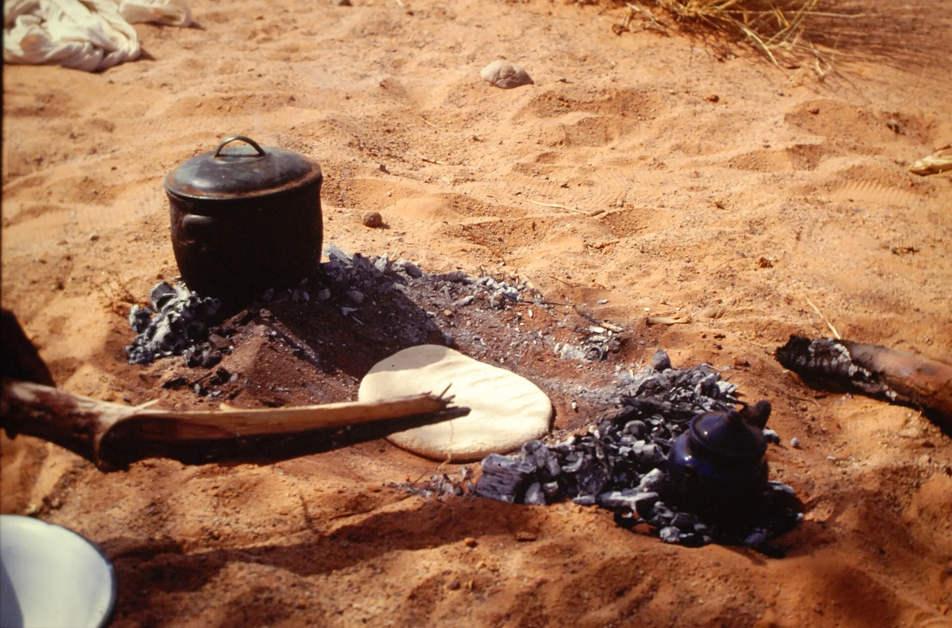 Sur le sable, une galette de pâte claire posée sur un lit de braises noircies, à côté d’une marmite en fonte noire ; un morceau de bois brûlé au premier plan