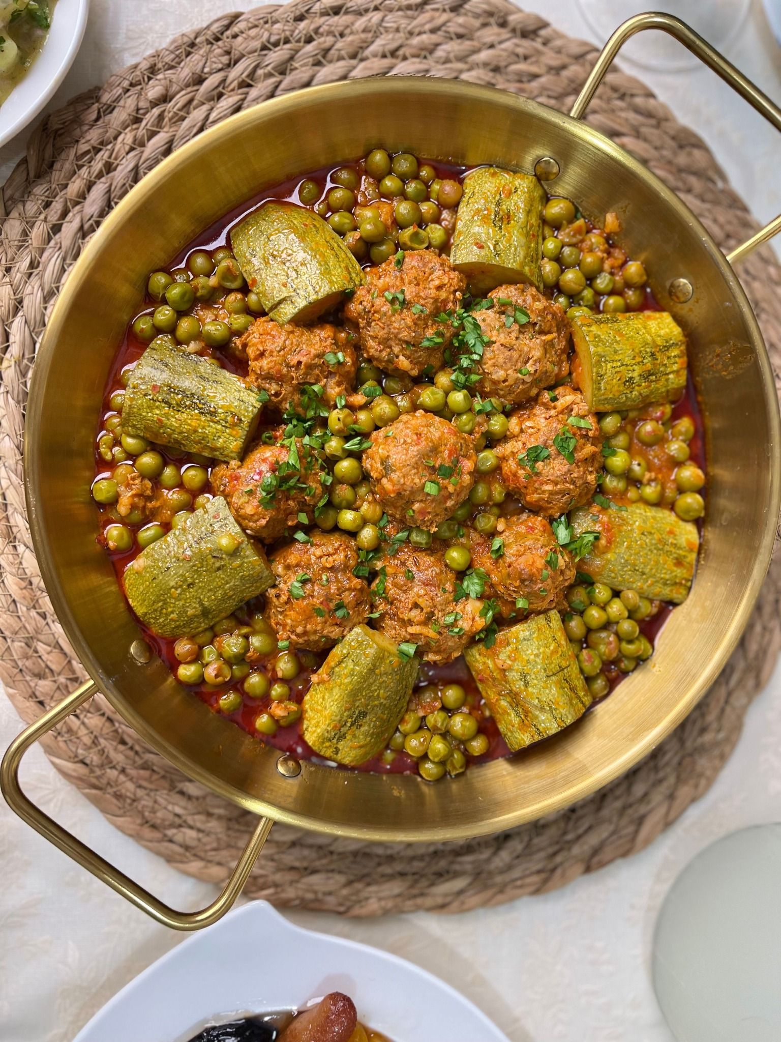 Vue du dessus d’un plat en laiton à deux anses, posé sur un set tressé : des boulettes, des petits pois et des tronçons de courgette dans une sauce rouge, parsemés d’herbes hachées