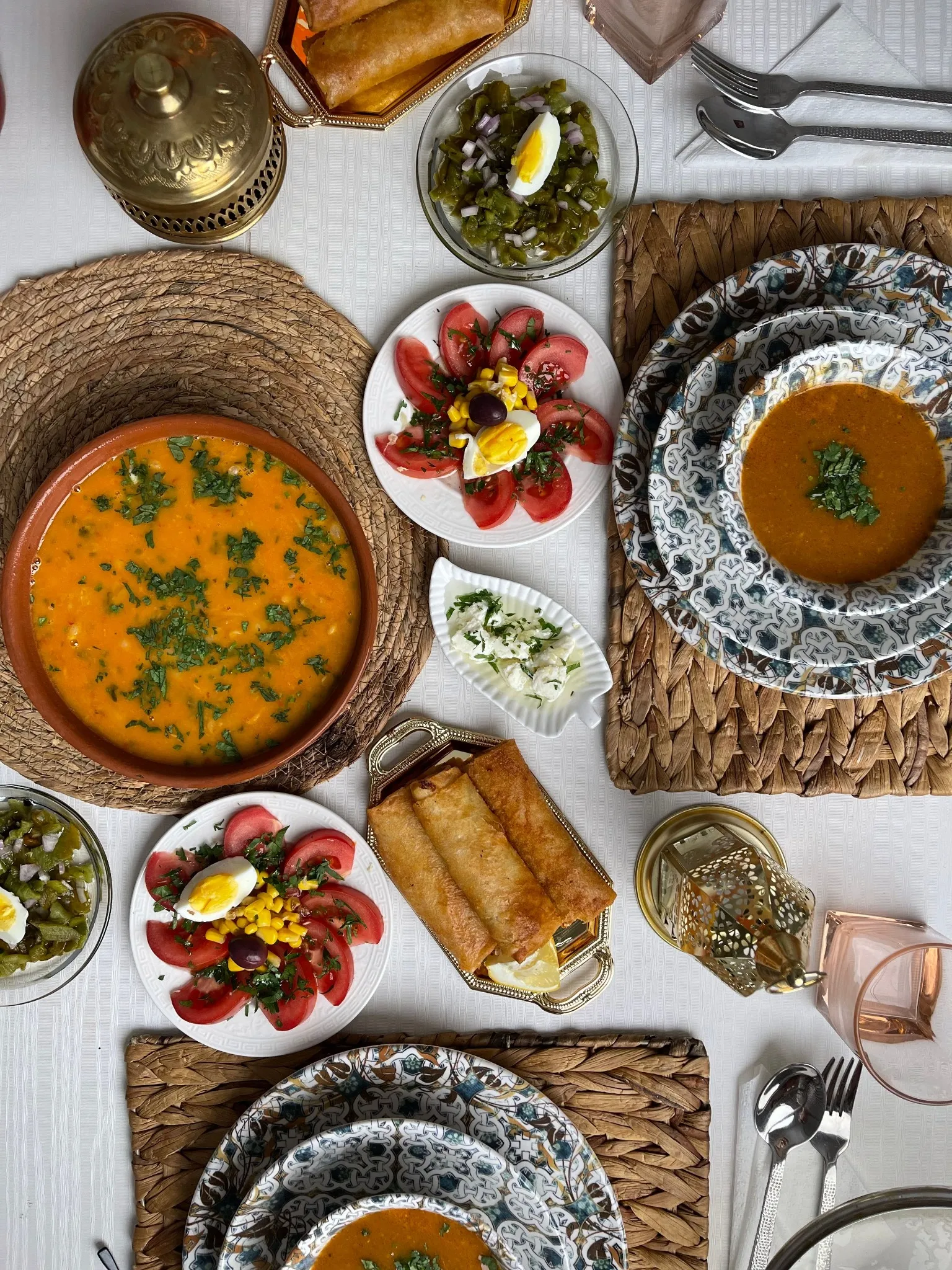 Table vue du dessus : une soupe orangée parsemée d’herbes dans un grand bol en terre, une autre soupe dans des assiettes à motifs bleus posées sur un set tressé, des assiettes de tomates garnies d’œuf dur, de maïs et d’olive, des feuilletés roulés et dorés, une petite lanterne et une boîte en laiton