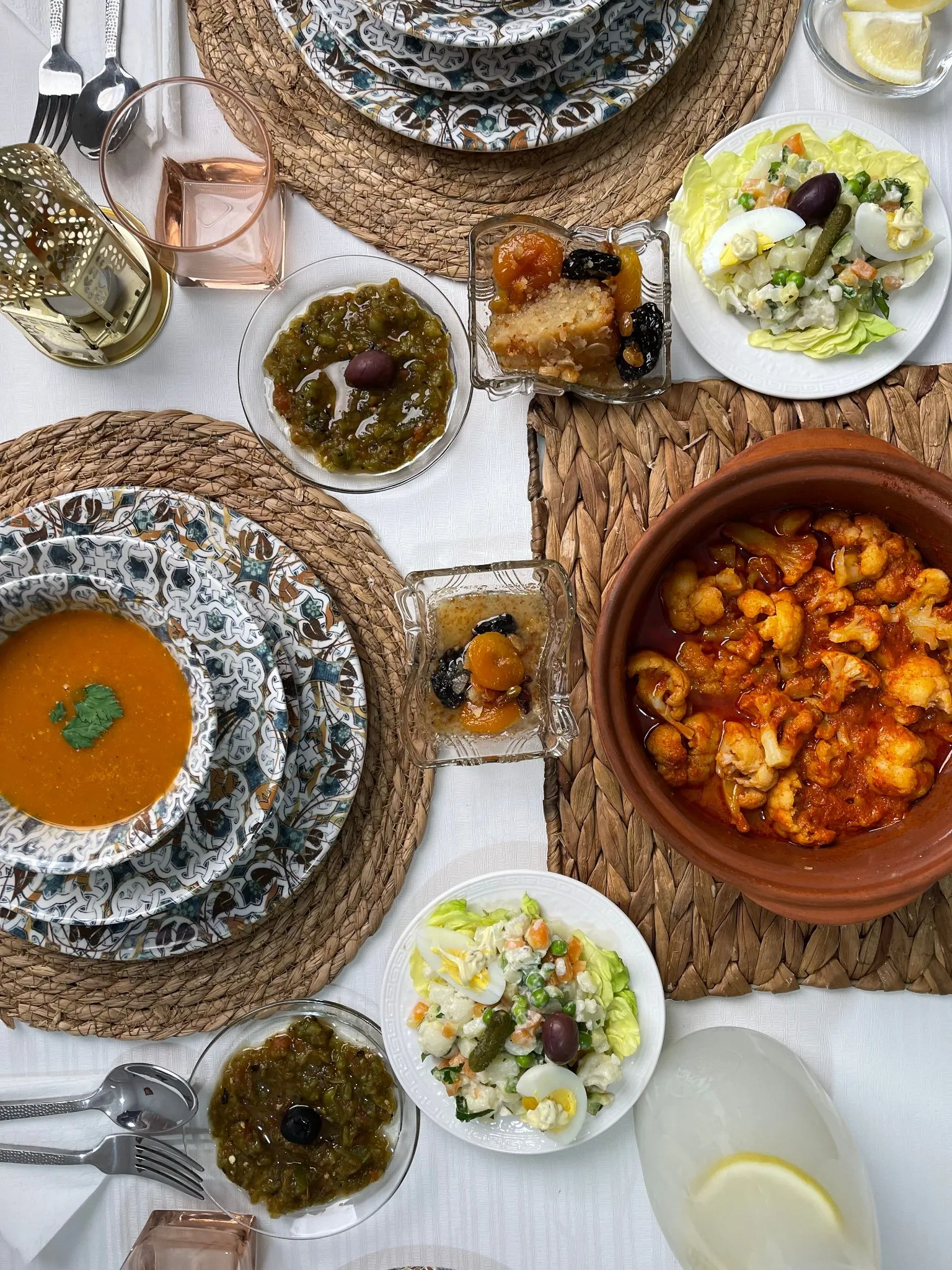 Table vue du dessus : à droite, une cocotte en terre pleine de bouquets de chou-fleur dans une sauce rouge ; à gauche, une soupe orangée dans des assiettes à motifs bleus ; entre les deux, des salades composées garnies d’œuf dur et d’olive, des coupelles de légumes en sauce et de petites coupes de pruneaux et d’abricots