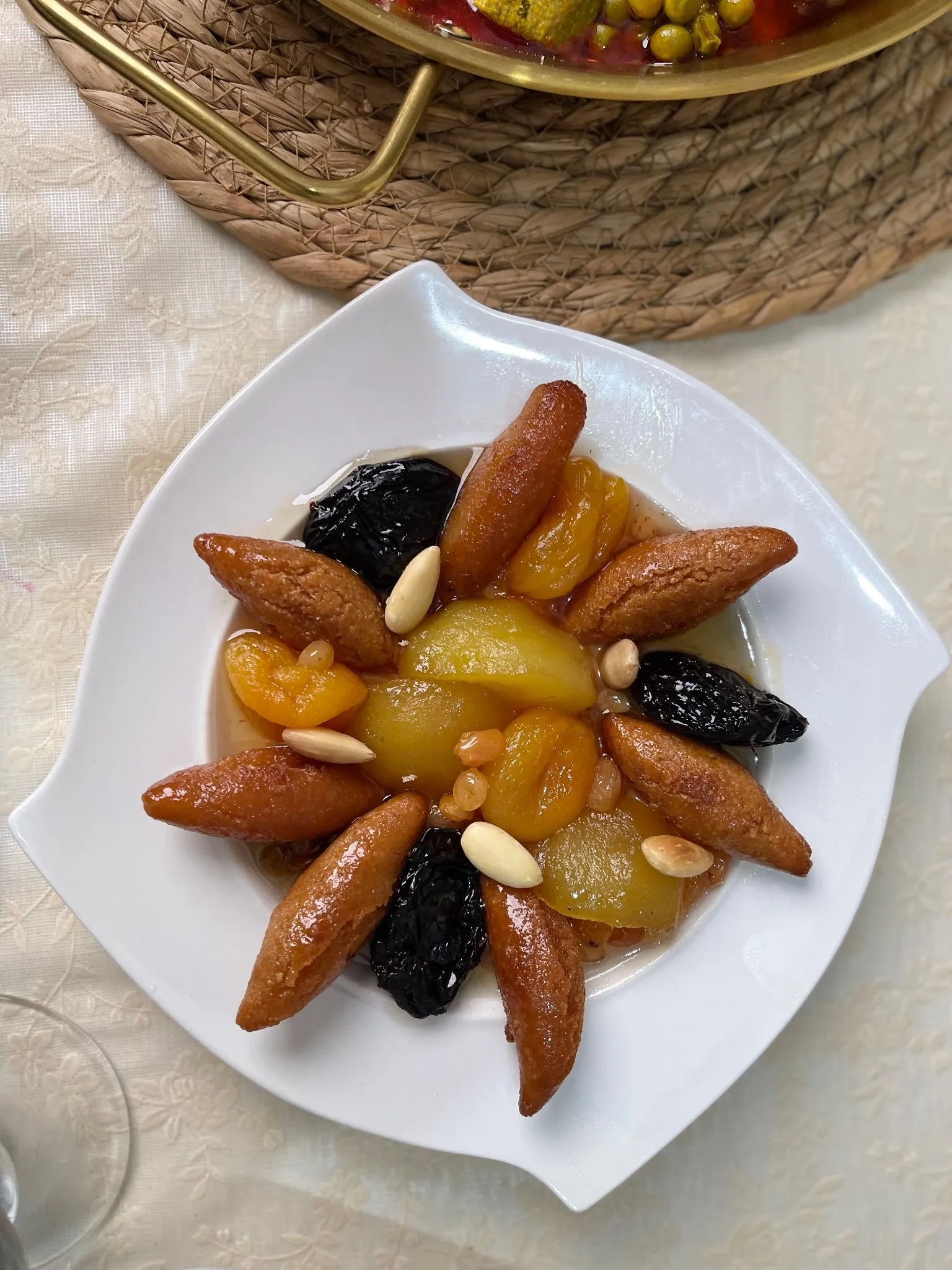 Vue du dessus d’une assiette blanche aux bords ondulés : des pruneaux, des abricots secs, des quartiers de fruit clair et des amandes dans un sirop ambré, entourés de pièces dorées en forme de fuseau disposées en étoile