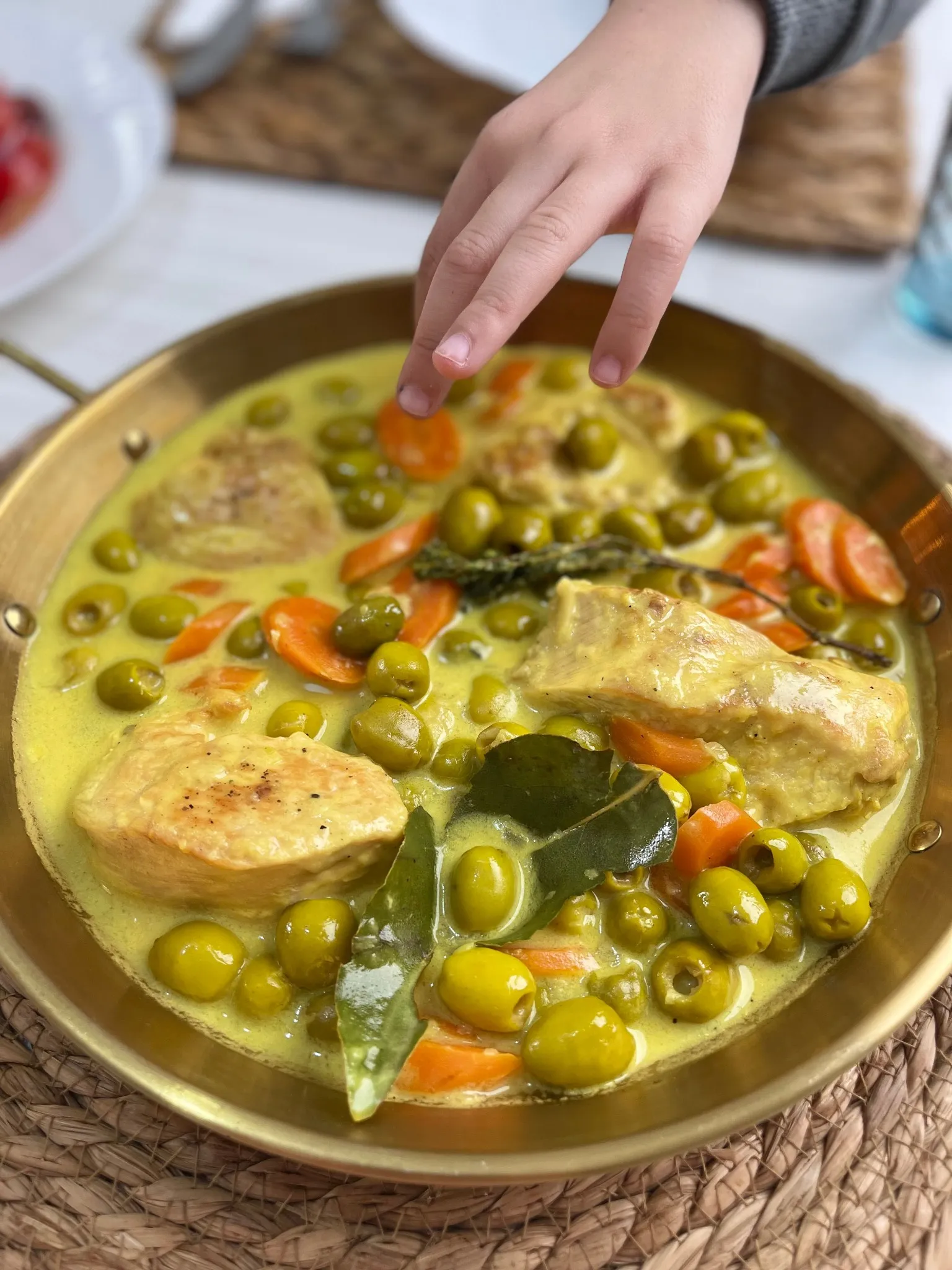 Un plat en laiton rempli d’une sauce jaune où baignent des olives vertes, des rondelles de carotte, des morceaux de viande dorés, des feuilles de laurier et un brin de thym ; une petite main s’approche pour y piocher