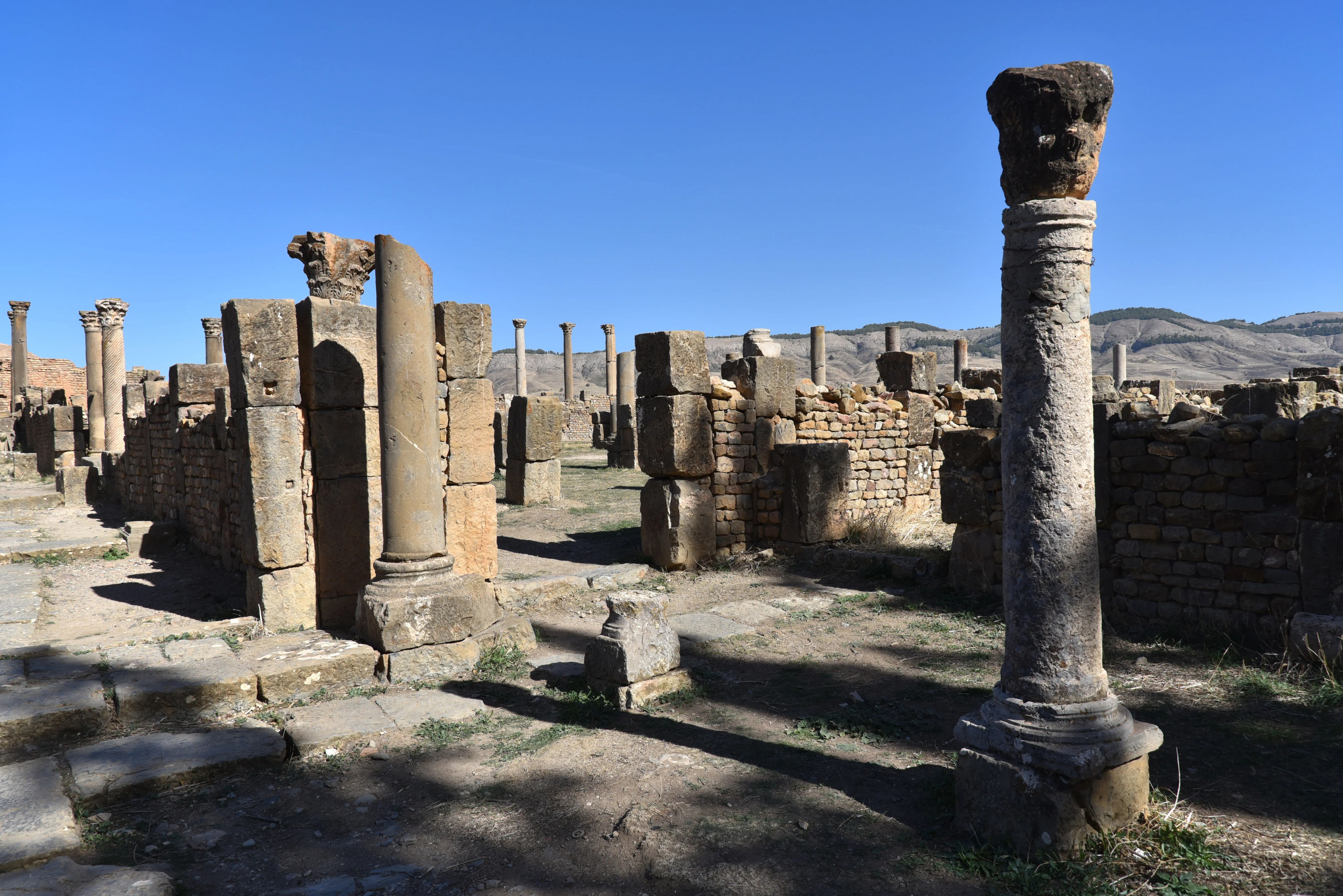 Rue dallée bordée de colonnes et de murs de pierre en partie écroulés ; au premier plan, une colonne isolée au chapiteau brisé, et au fond des collines sèches sous un ciel bleu