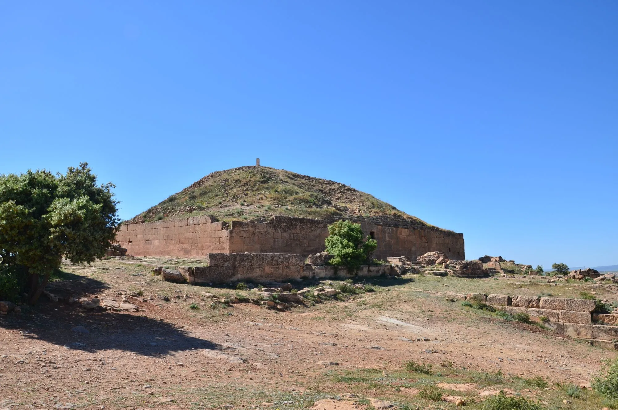 Mausolée en pierre ocre à base carrée, surmonté d’un tertre pyramidal couvert d’herbe, sur une colline sèche sous un ciel bleu ; un arbuste au premier plan à gauche et des blocs épars autour