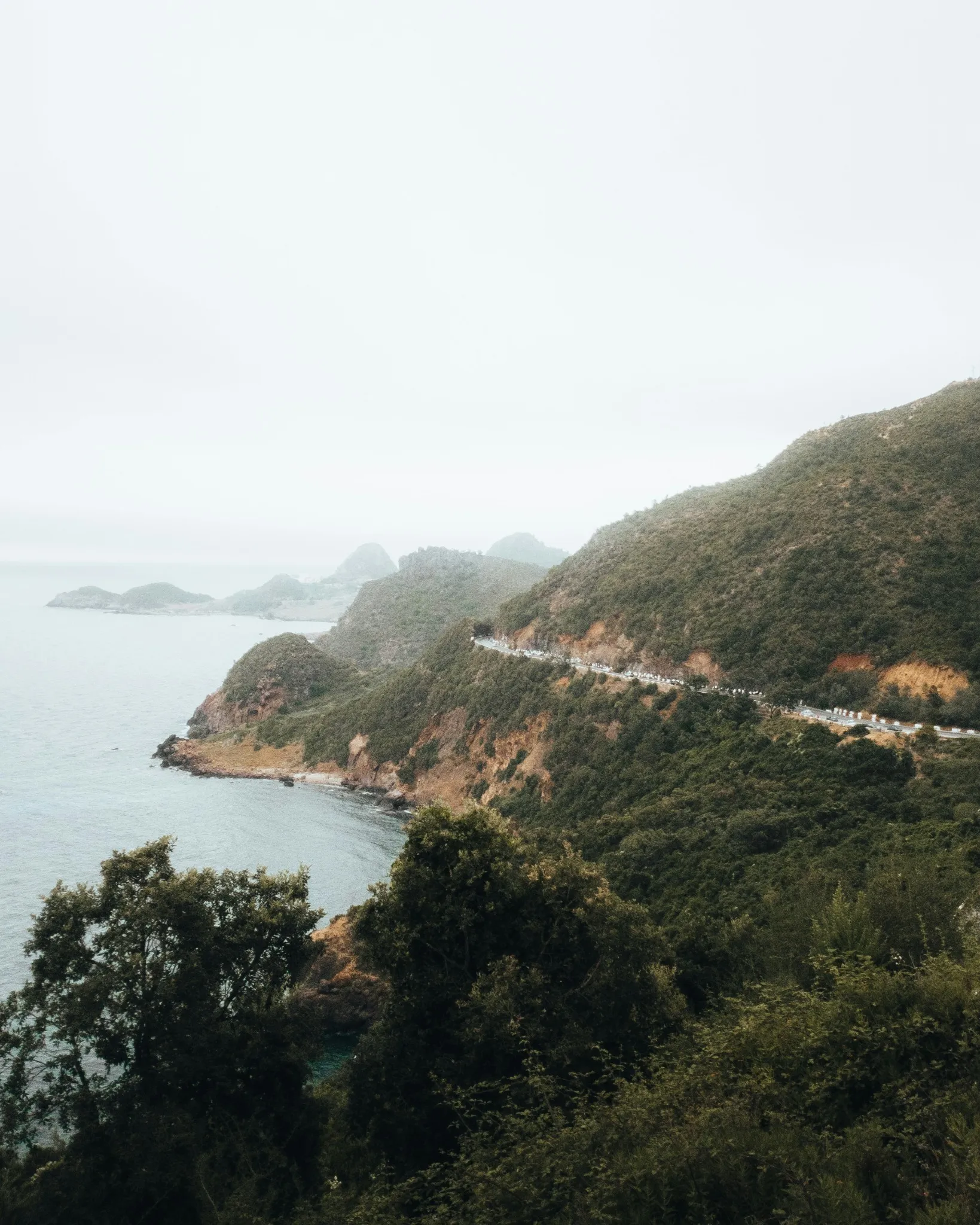 Collines couvertes de maquis qui plongent dans une mer grise ; une route taillée à flanc de falaise longe la côte, et des îlots se perdent dans la brume au loin