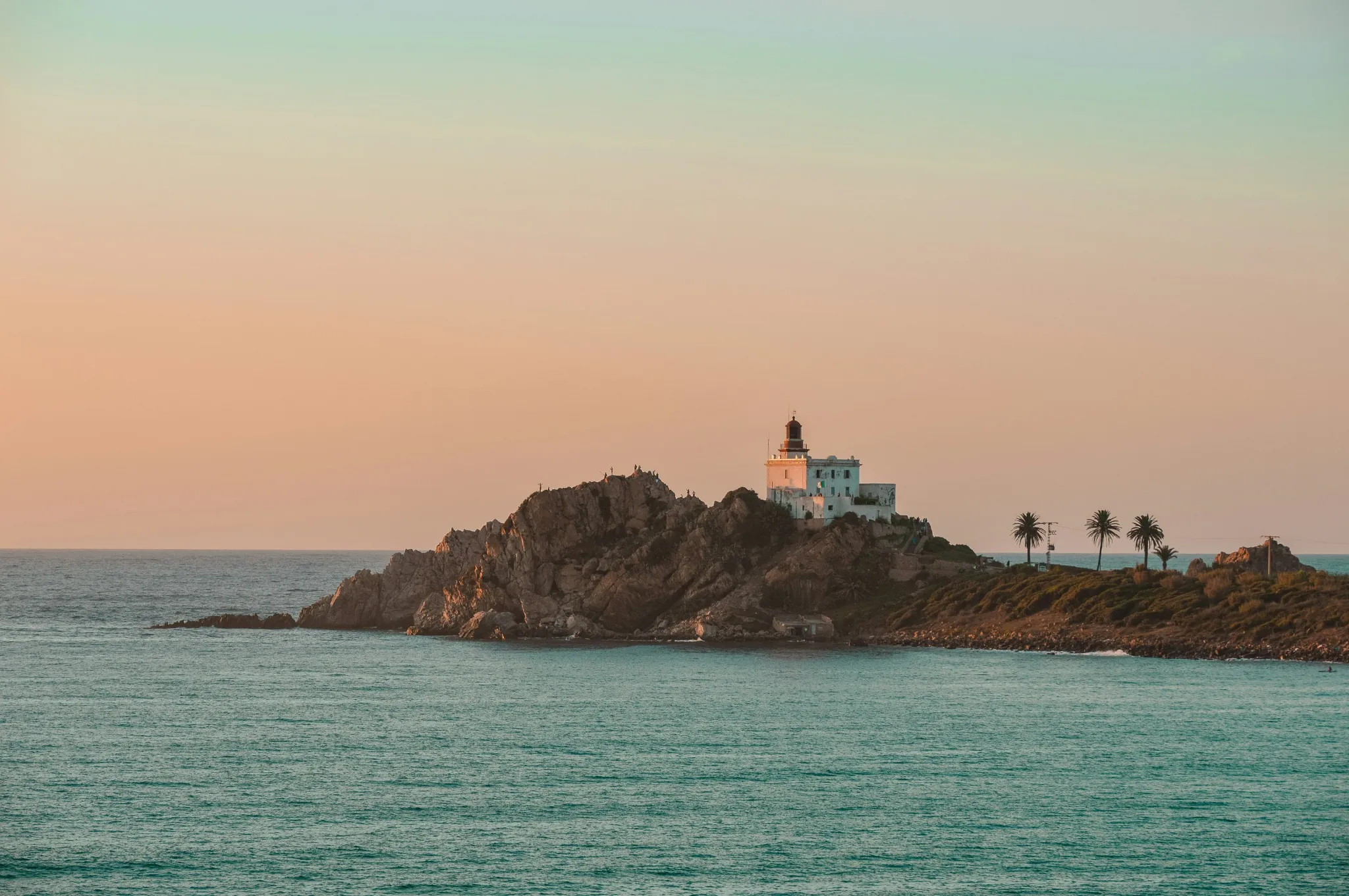 Un phare blanc, tour et bâtiment, posé sur un promontoire rocheux au-dessus d’une mer vert d’eau ; quelques palmiers à droite, sous un ciel rosé