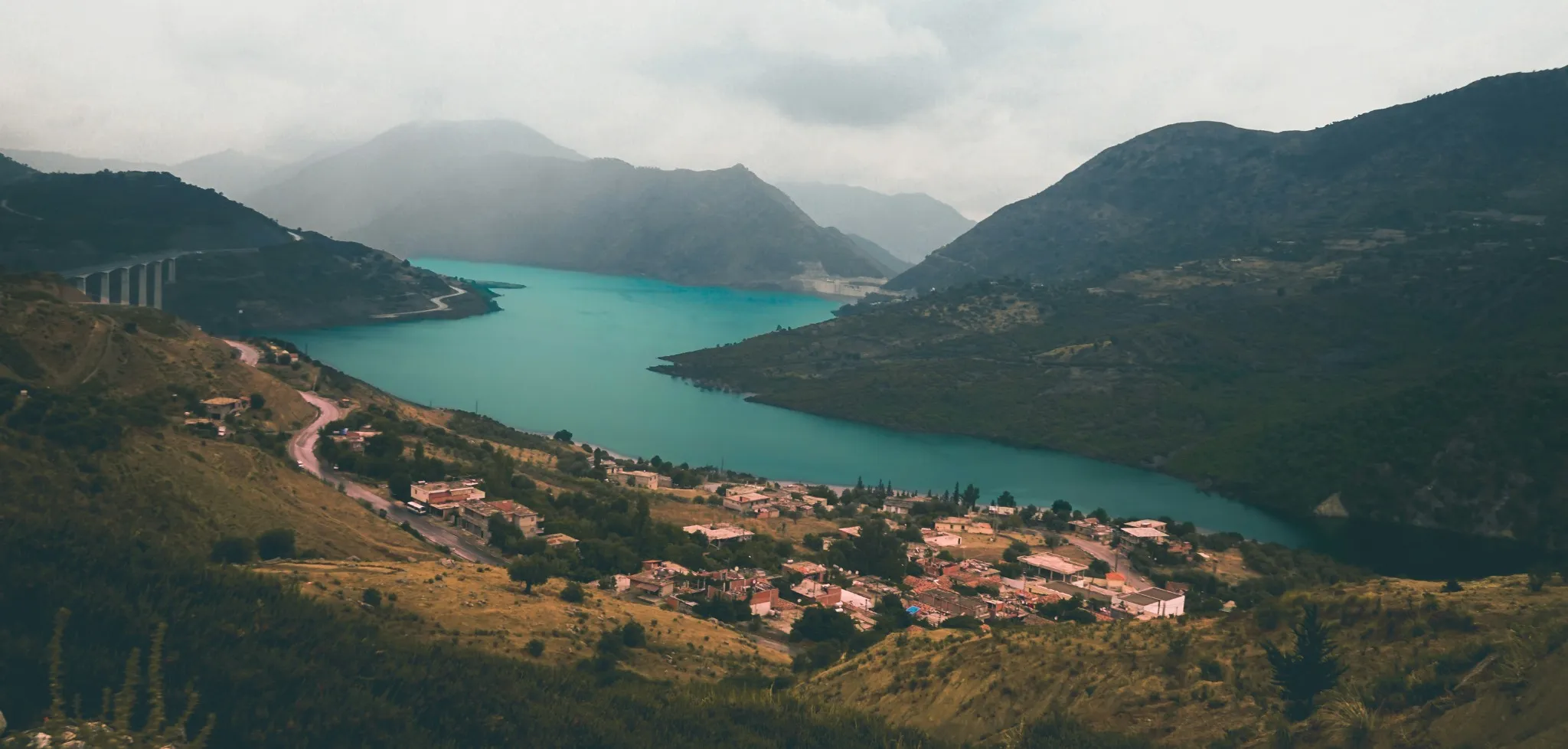 Vue d’en haut sur un lac turquoise entre des montagnes sous un ciel couvert ; un viaduc le franchit à gauche et un village aux toits roses s’étire sur la rive, au bord d’une route en lacets