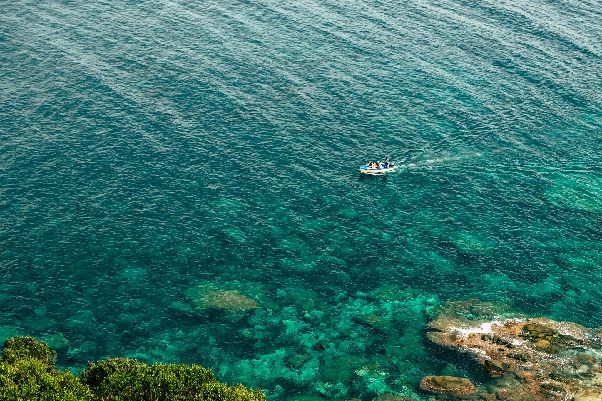 Vue plongeante sur une mer turquoise si claire que l’on devine les rochers du fond ; une petite barque blanche y trace son sillage, au-dessus de rochers et de buissons au premier plan