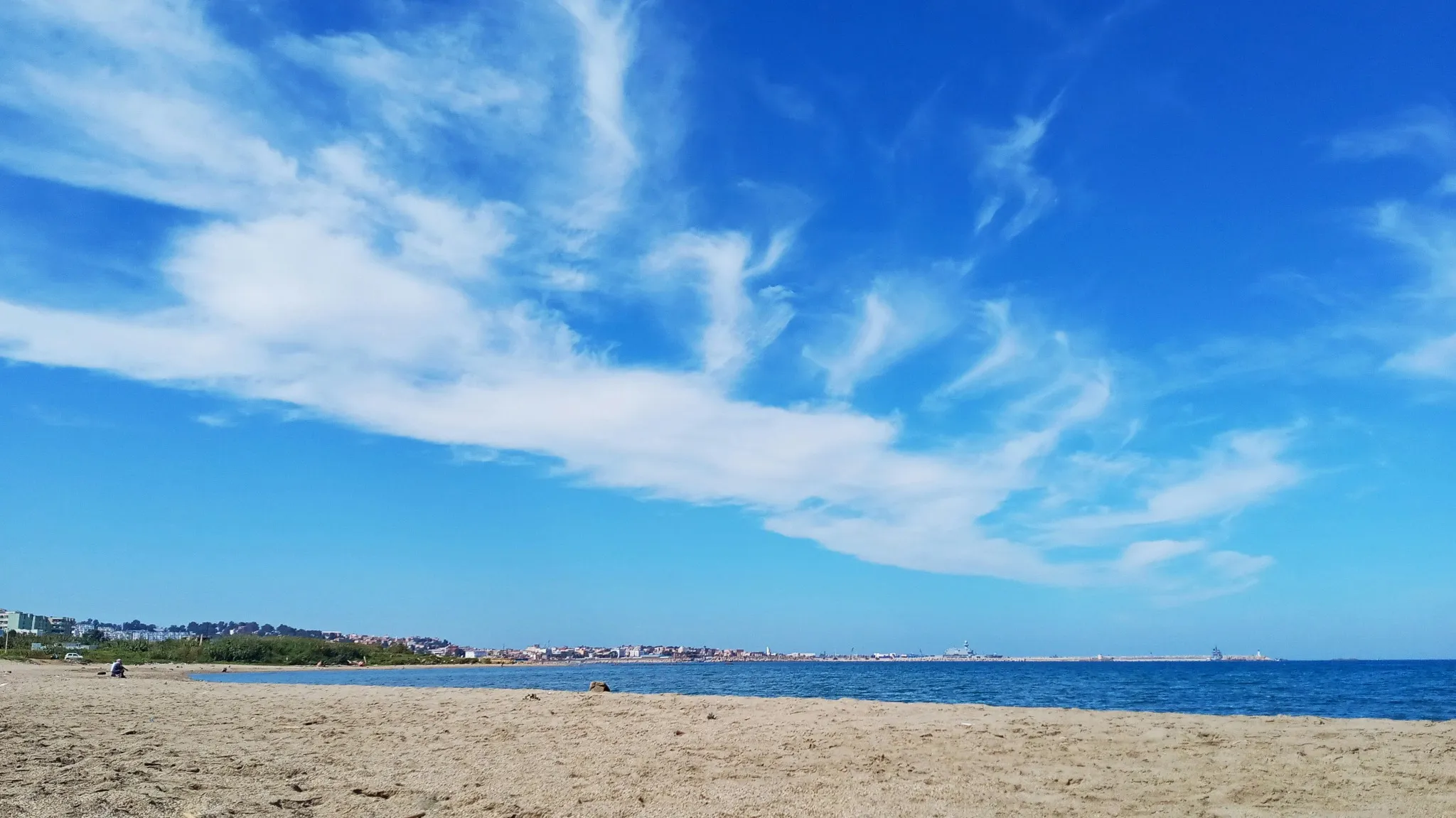 Grande plage de sable sous un ciel bleu traversé de longs nuages blancs ; au loin, une ville basse et une jetée bordent la mer