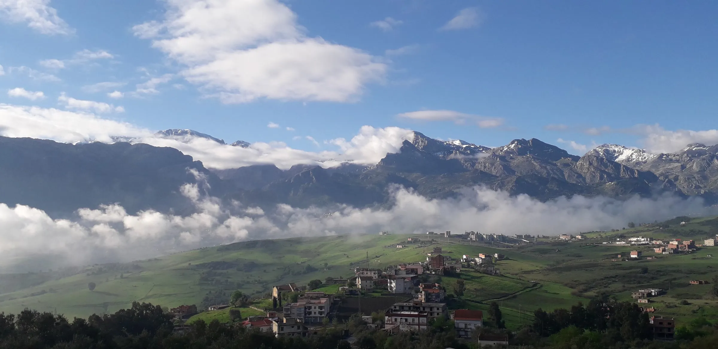 Un village de maisons blanches dispersées dans des prés verts ; derrière, une longue chaîne de montagnes enneigées émerge d’une mer de nuages sous un ciel bleu