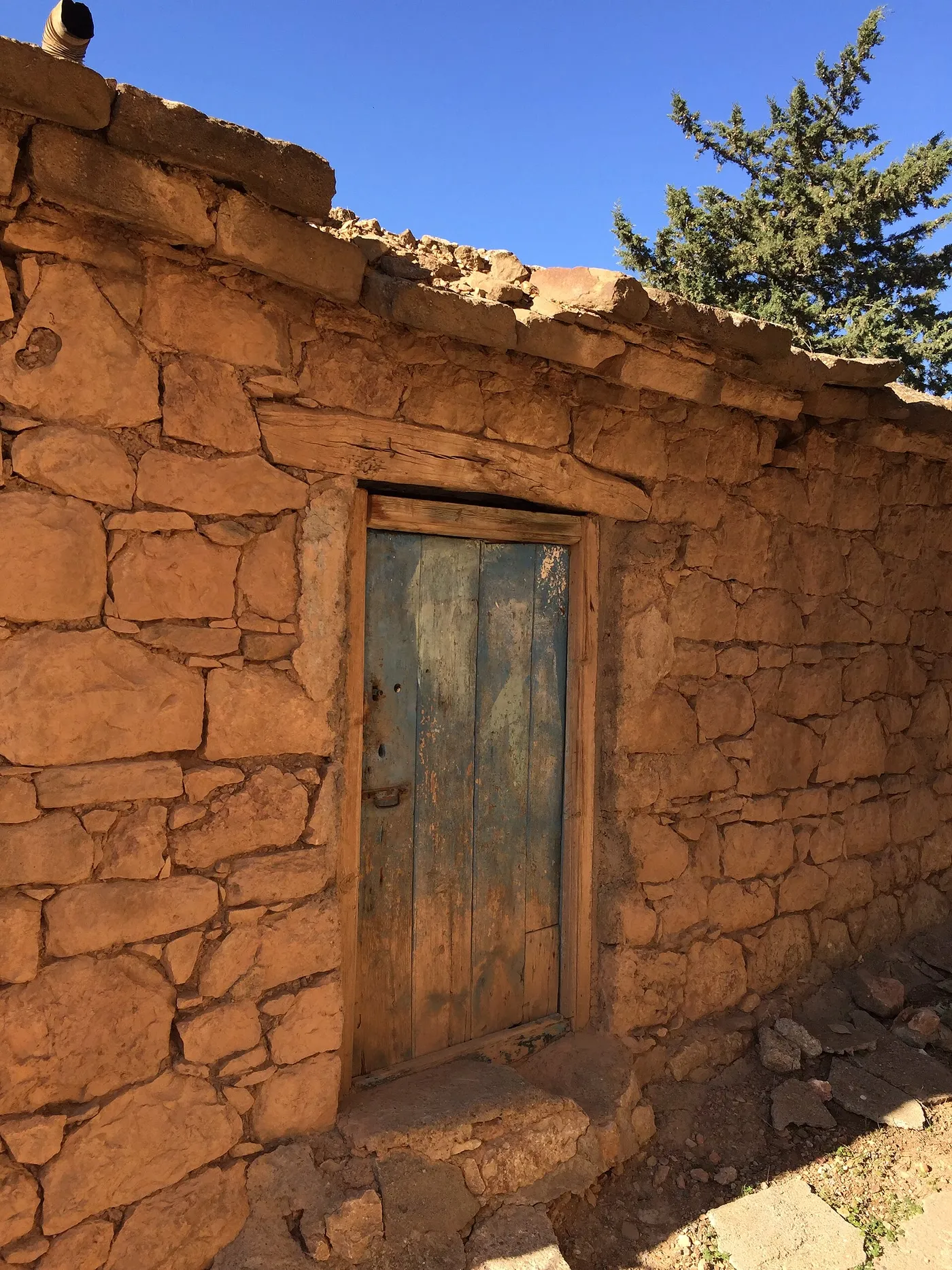 Une maison basse en pierres ocre assemblées sans enduit, percée d’une porte de bois patinée, sous un ciel bleu