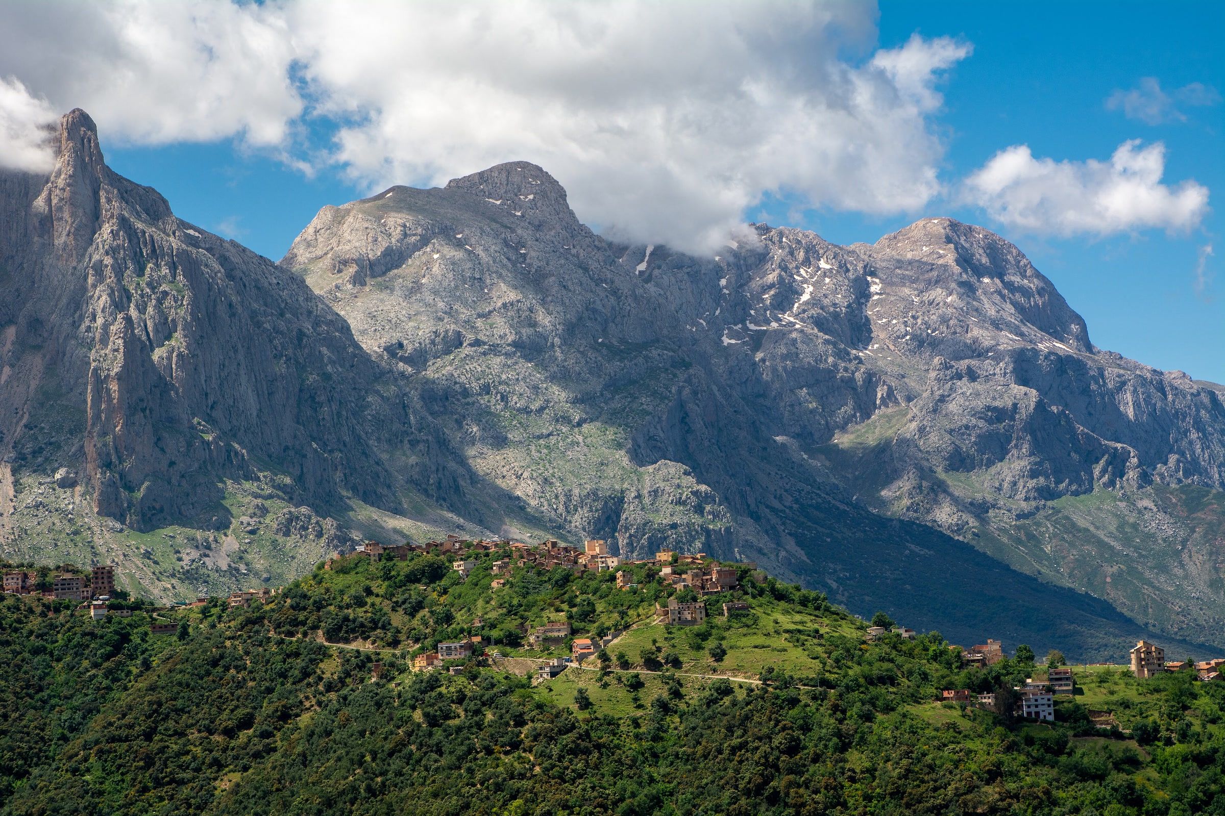 De hautes falaises calcaires grises, striées de quelques névés, dominent une crête verte où s’égrènent les maisons d’un village, sous un ciel bleu et de gros nuages blancs
