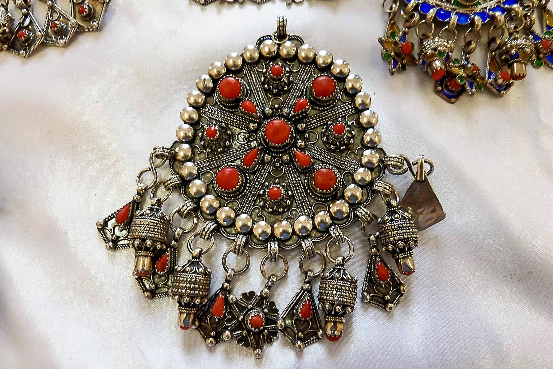 Gros plan sur un pendentif rond en argent, orné de perles d’argent et de cabochons de corail rouge, d’où pendent de petites pièces émaillées