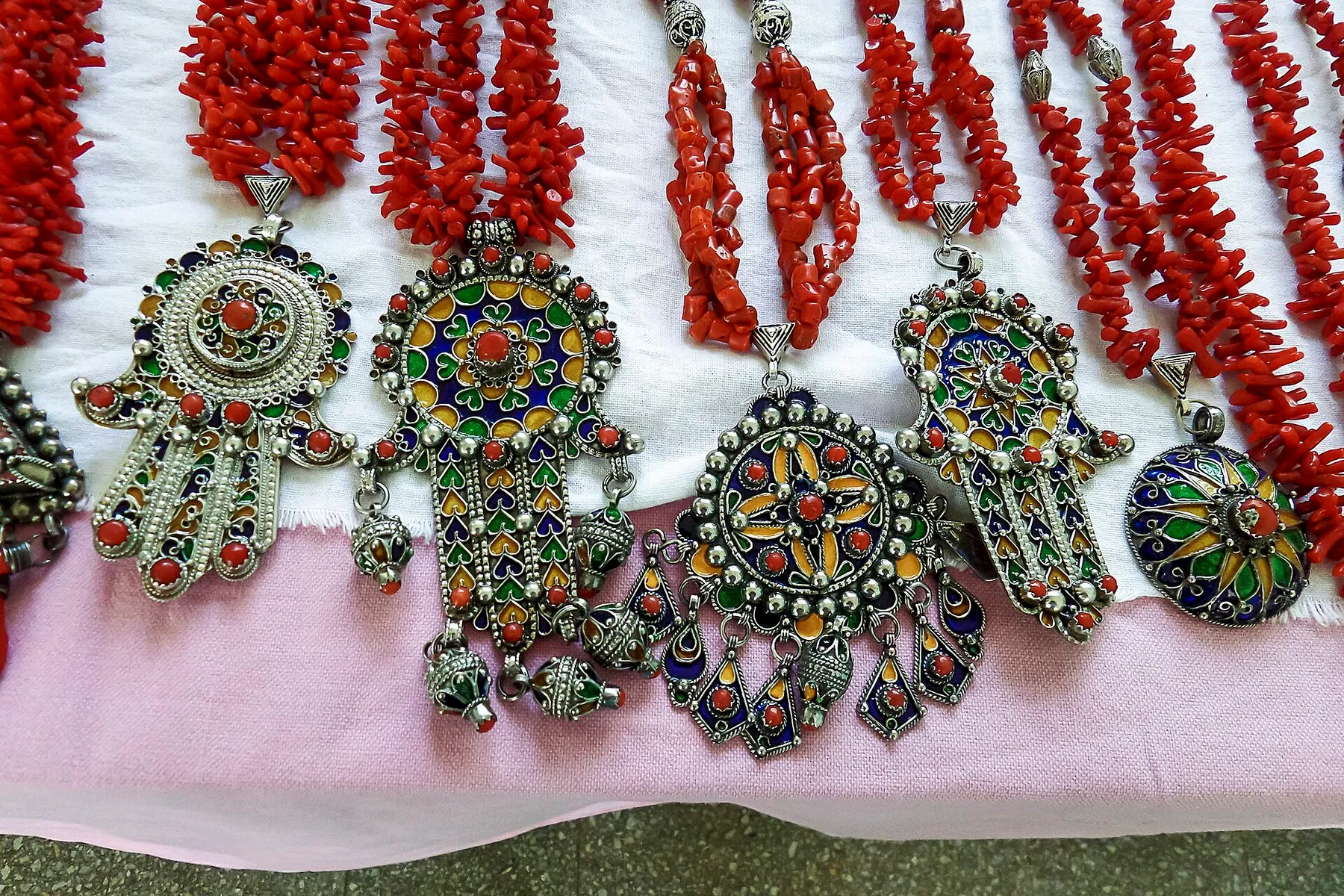 Des pendentifs en argent émaillés de bleu, de vert et de jaune, en forme de main ou de disque, sertis de corail et suspendus à des colliers de branches de corail rouge, posés sur un tissu clair