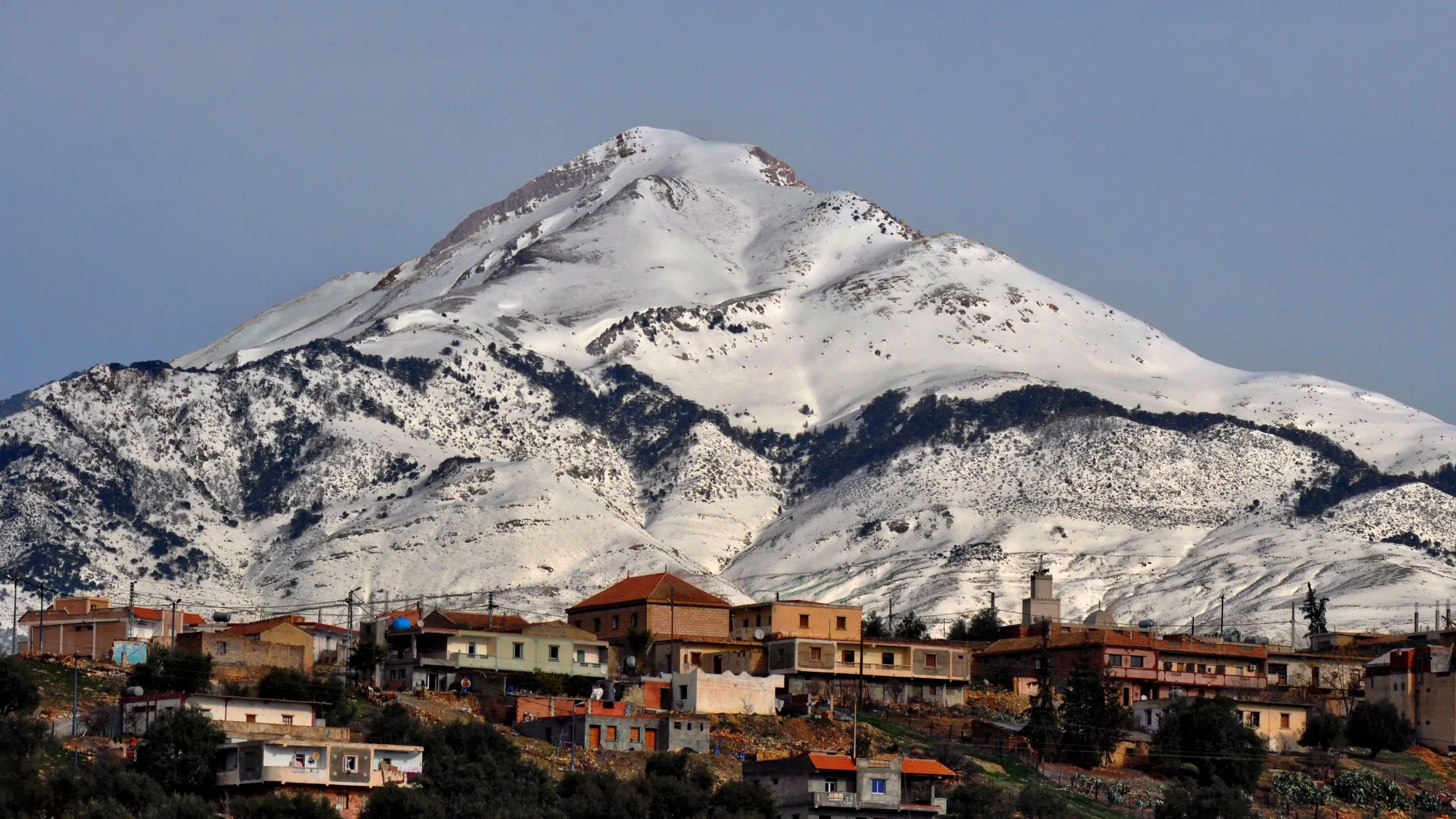 Un sommet massif entièrement enneigé domine un village de maisons aux toits rouges, au premier plan, sous un ciel gris-bleu