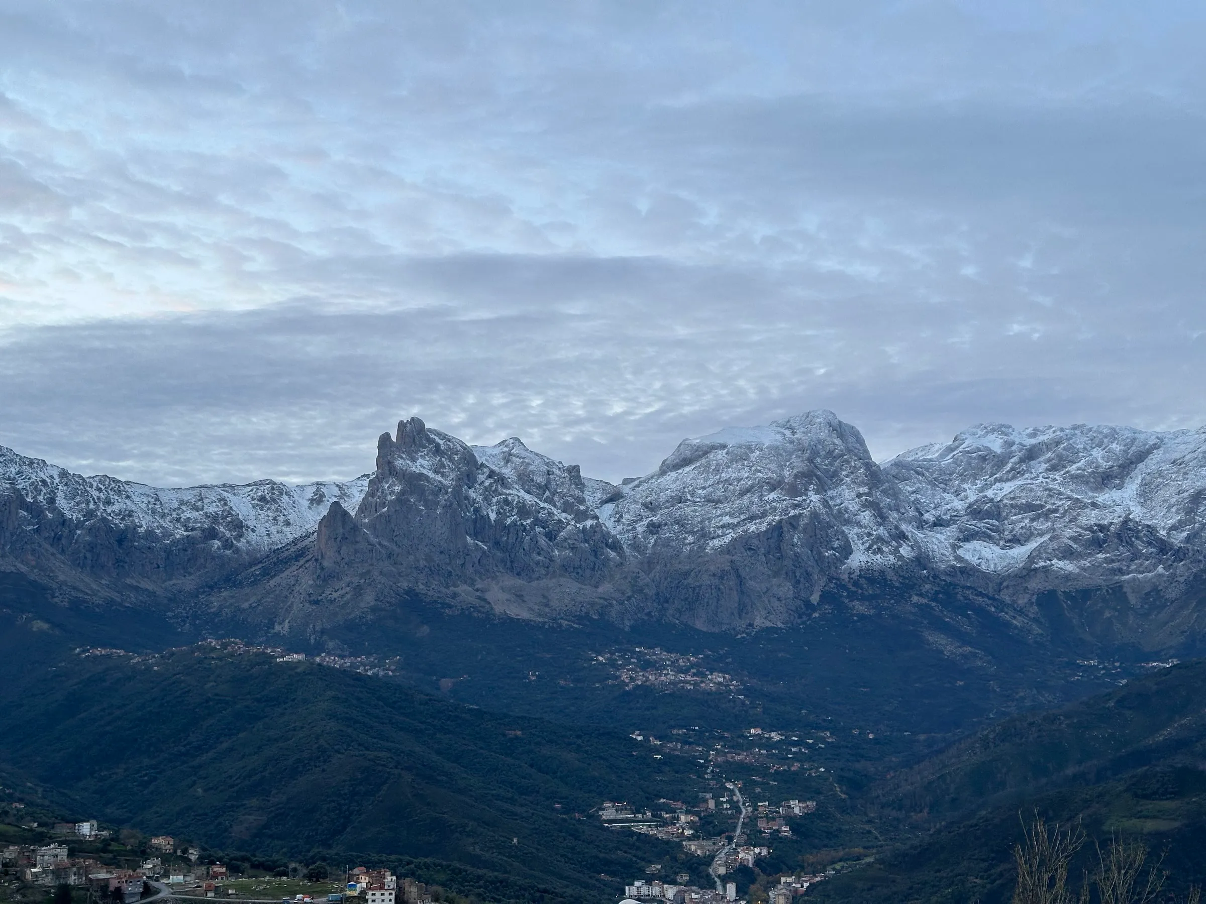 Une chaîne de montagnes aux sommets déchiquetés, saupoudrés de neige, au-dessus d’une vallée sombre où l’on devine des villages ; le ciel est couvert d’un voile de nuages au lever du jour