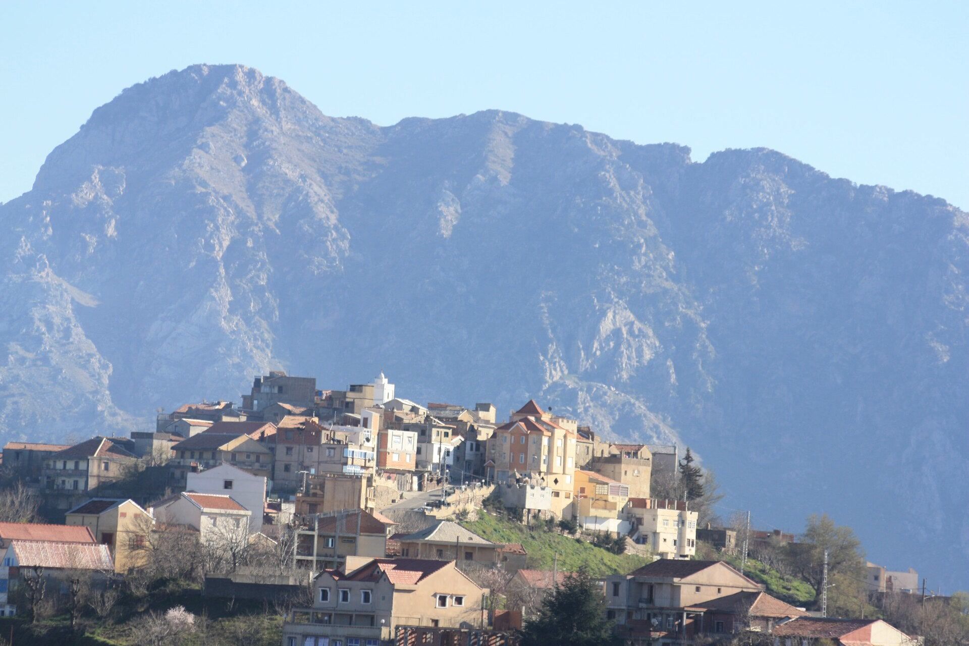 Un village dense de maisons claires aux toits de tuiles, étagé sur une colline, au pied d’une immense paroi rocheuse bleutée dans la lumière du matin