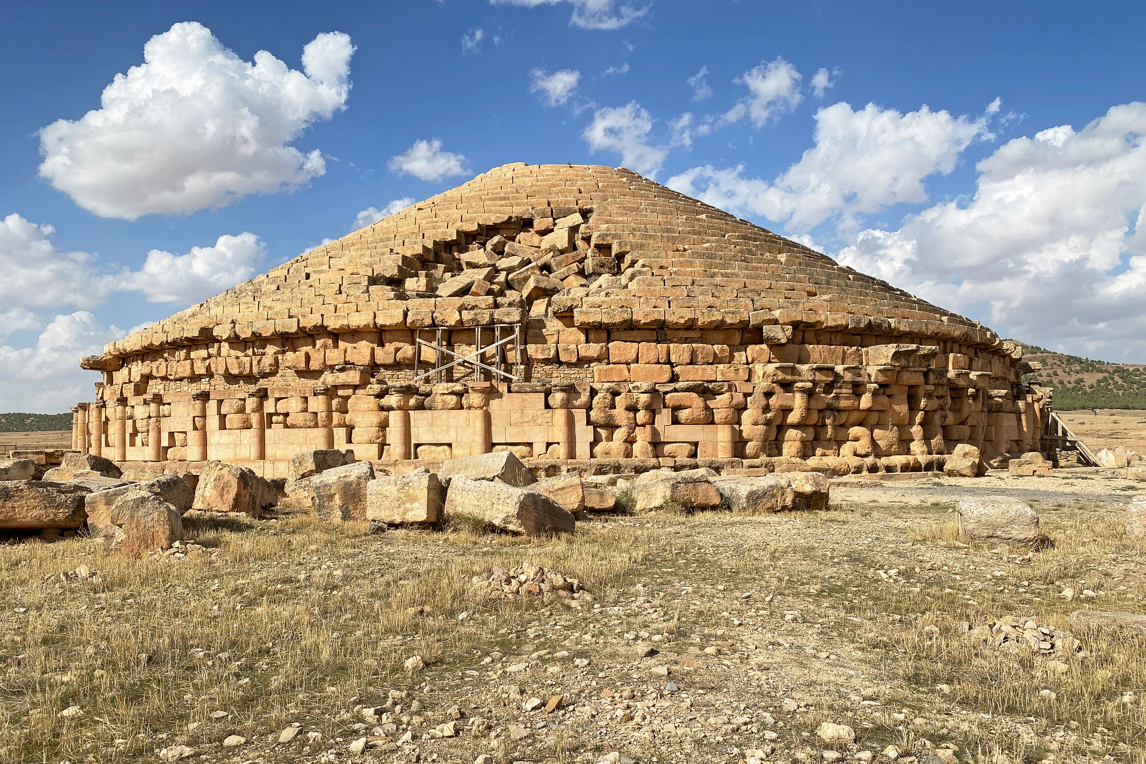 Grand monument circulaire en pierre ocre, entouré de colonnes engagées et coiffé de gradins en cône, dont une partie est effondrée ; un échafaudage métallique est posé contre la façade, des blocs épars jonchent la steppe au premier plan