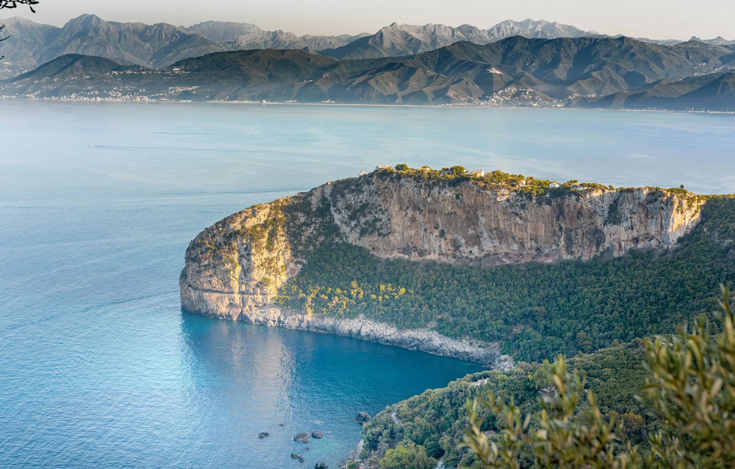 Vue d’en haut sur un cap couvert de forêt, bordé de falaises claires, qui s’avance dans une mer calme ; au fond, une longue côte et des montagnes