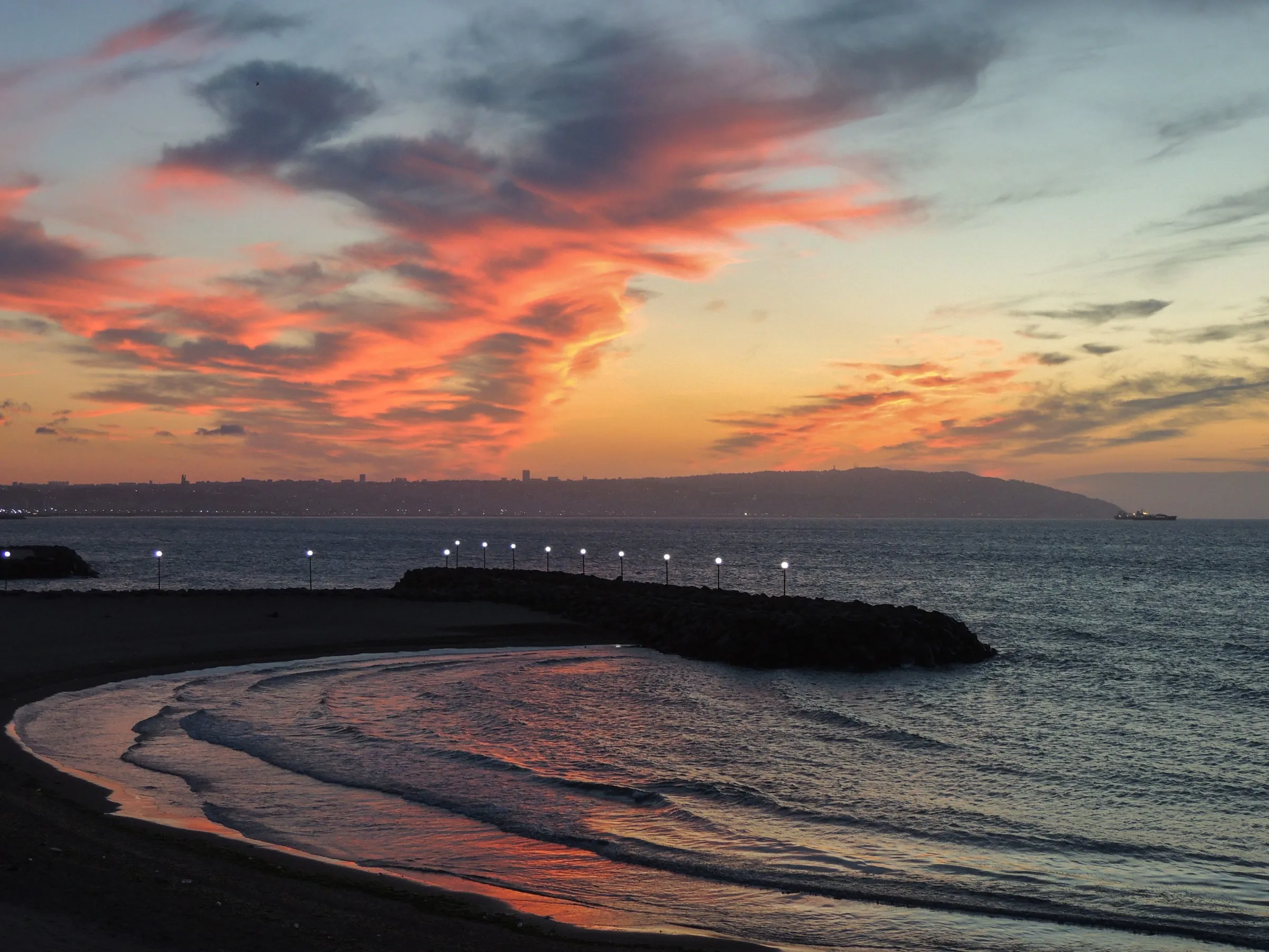Crépuscule sur une baie : une plage en arc de cercle, une jetée éclairée de lampadaires et, au loin, une ville sur des collines, sous un ciel strié de nuages roses