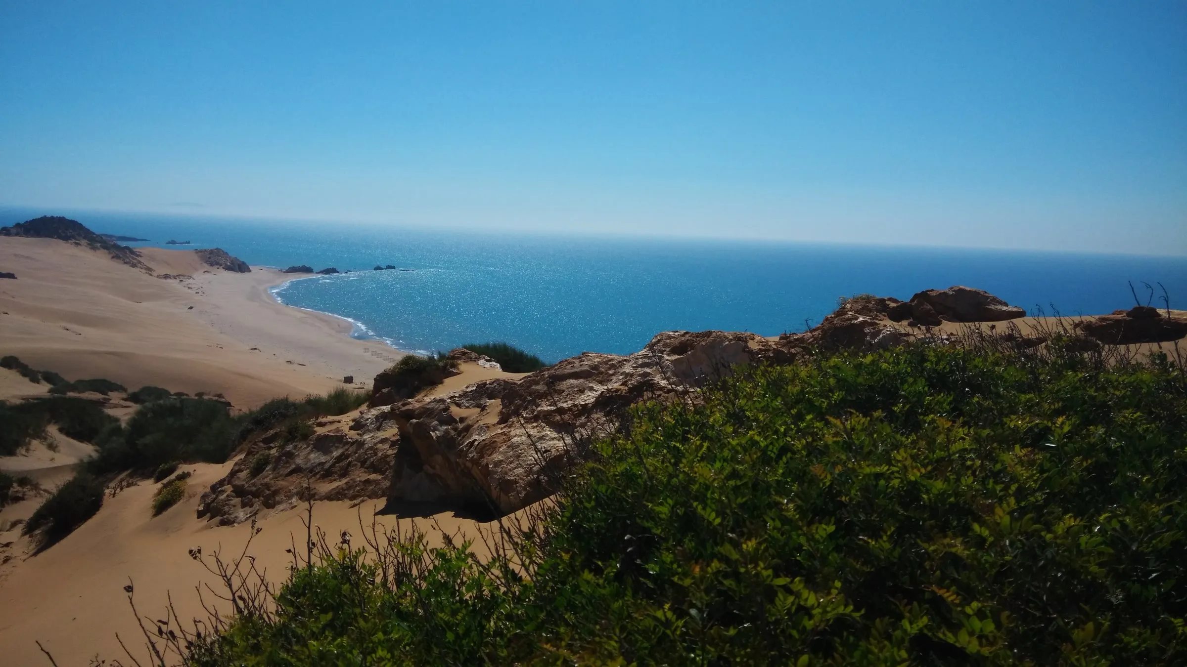 Des dunes de sable clair et des buissons descendent vers une longue plage et une mer d’un bleu profond ; au premier plan, des rochers et des broussailles