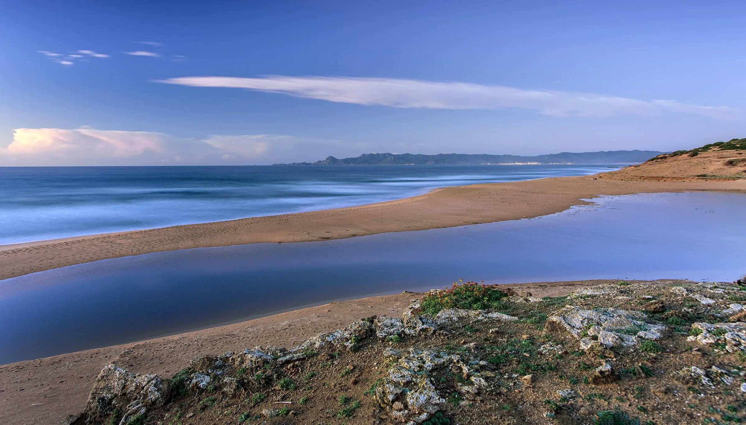Une langue de sable sépare la mer, aux vagues lissées par une longue pose, d’une lagune immobile ; au premier plan, des rochers couverts de lichen ; au loin, un cap
