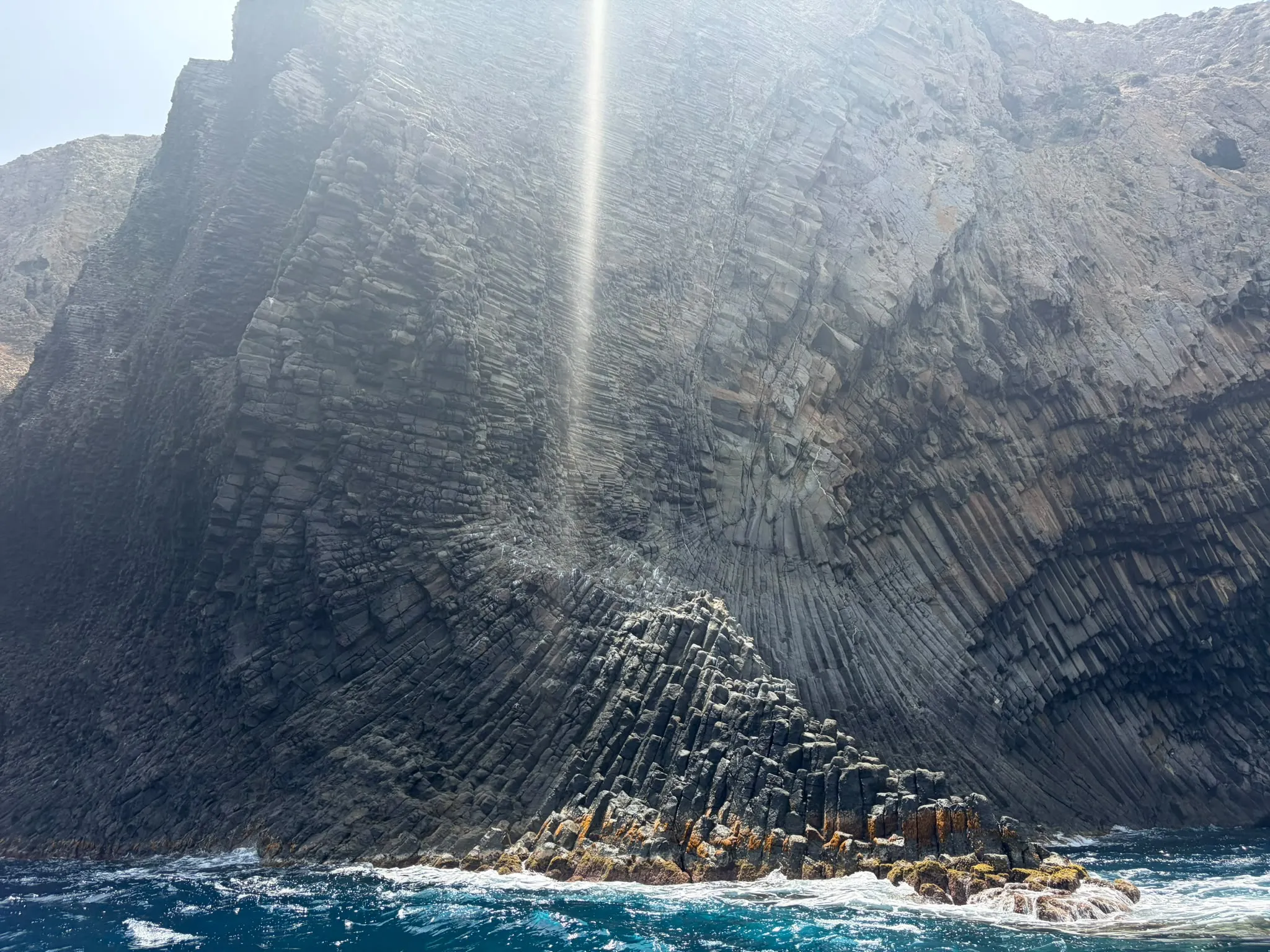 Une haute falaise sombre, striée de colonnes de roche dont certaines s’ouvrent en éventail, plonge dans une mer bleu profond frangée d’écume ; un voile de lumière et un reflet vertical traversent le haut de l’image, sous un coin de ciel clair à gauche