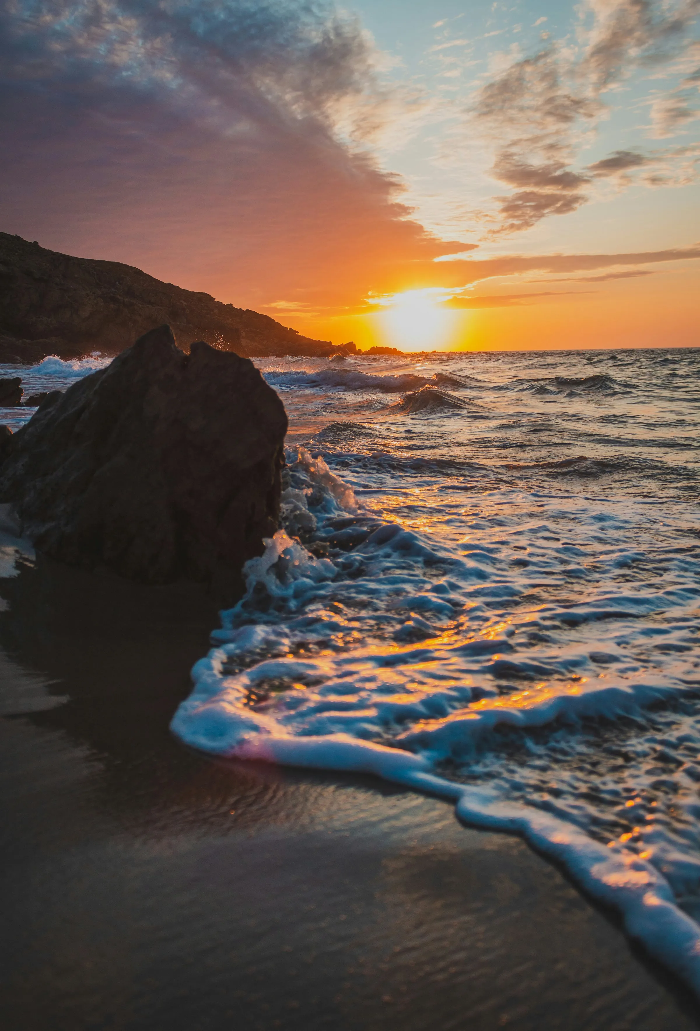 Au coucher du soleil, l’écume d’une vague s’étale sur le sable mouillé au pied d’un rocher sombre ; au loin, un cap et un ciel orangé