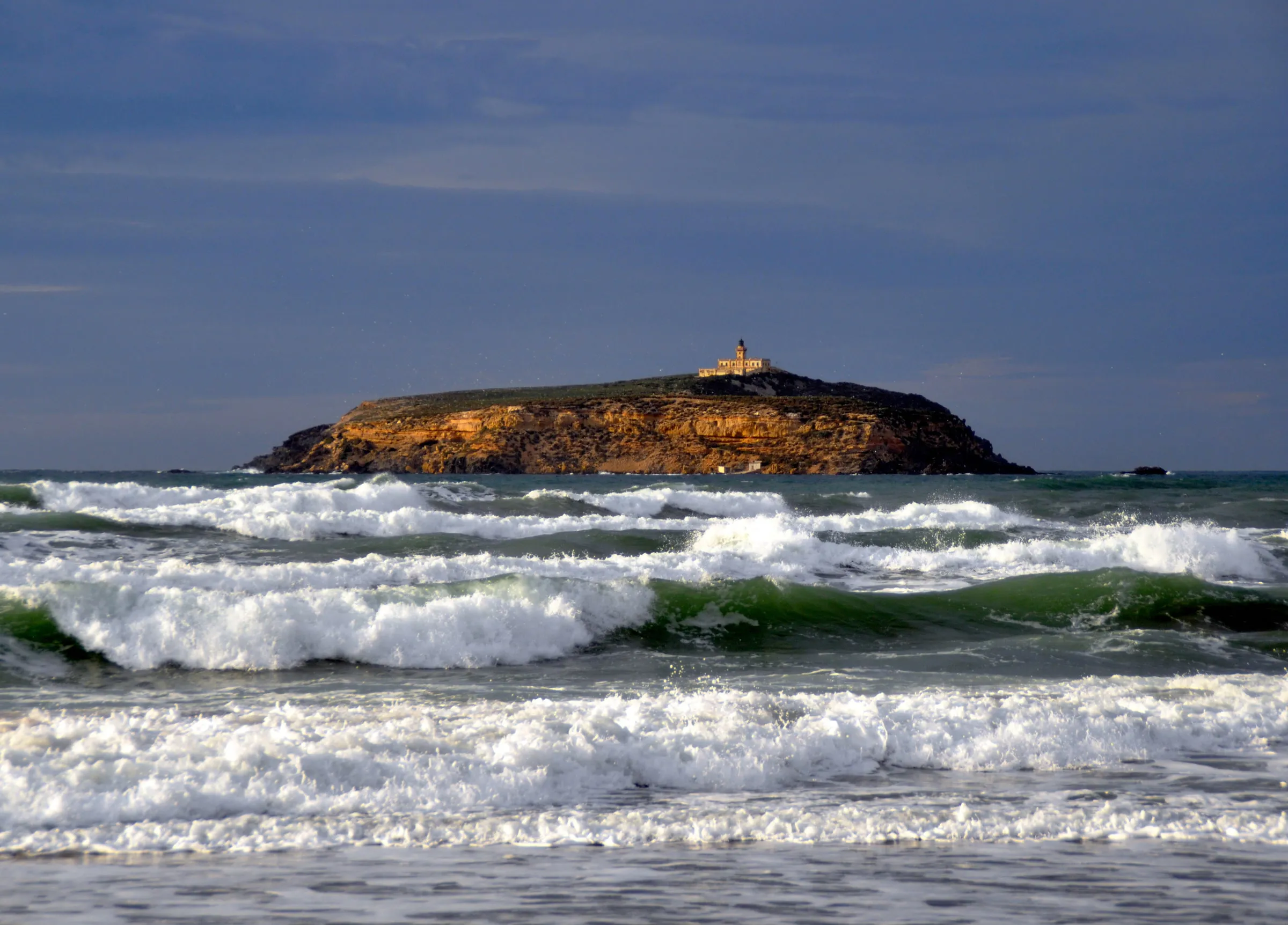 Une île plate aux falaises ocre, surmontée d’un phare, au large d’une mer agitée de rouleaux d’écume, sous un ciel d’orage
