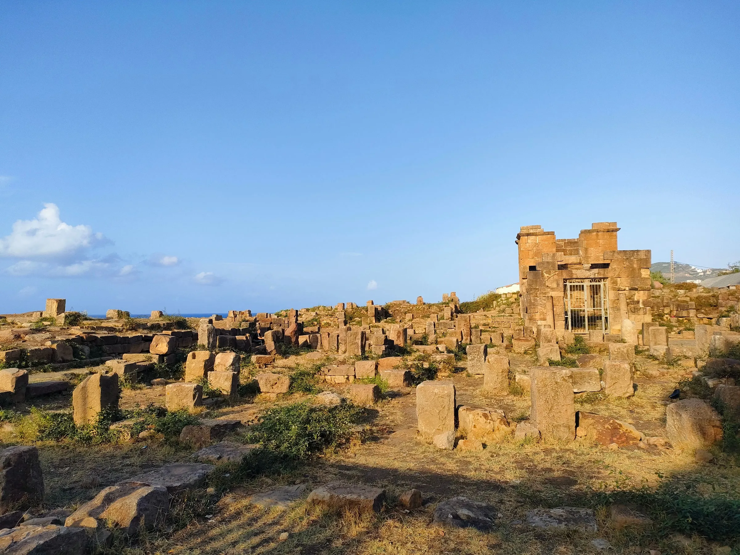 Un champ de blocs de pierre romains baignés par le soleil du soir, dominé par un petit édifice carré fermé d’une grille ; à gauche, un mince horizon de mer